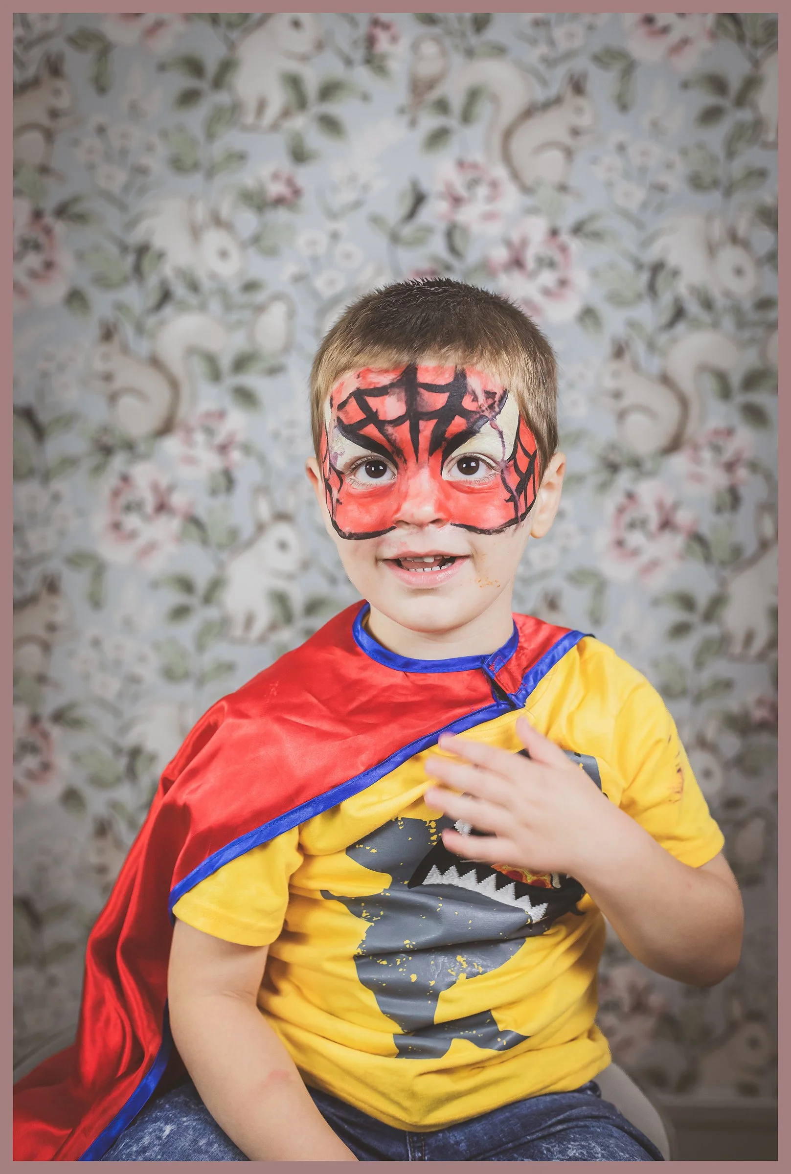 A young boy with face paint resembling Spider-Man's mask, wearing a yellow T-shirt with a dinosaur graphic, and a red cape with blue trim. He is smiling and touching his chest, sitting in front of a floral-patterned background.