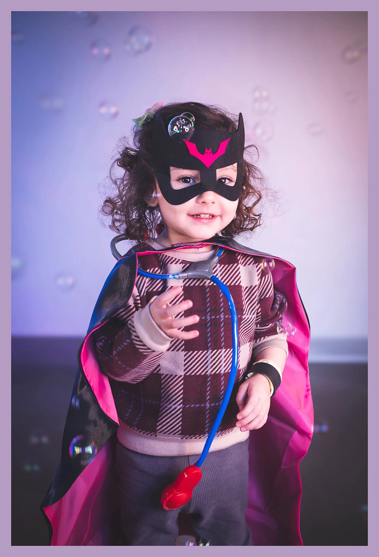 A young girl wearing a superhero mask with a pink bat symbol, a plaid sweater, and a dark cape with pink lining, holding a toy stethoscope and surrounded by floating bubbles.
