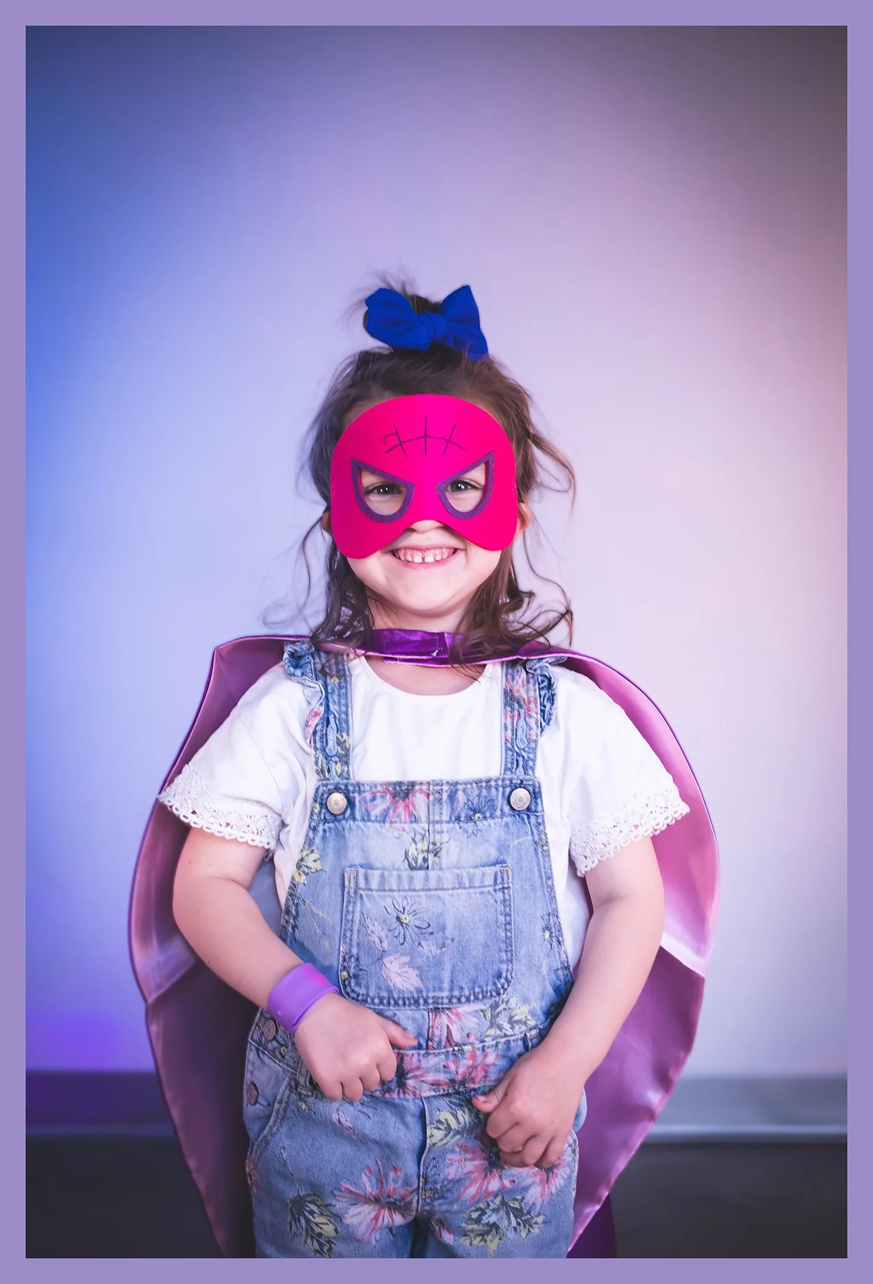 A young girl dressed as a superhero with a pink mask, a blue cape, a white top, denim overalls with floral print, and a blue bow in her hair, standing against a plain background.