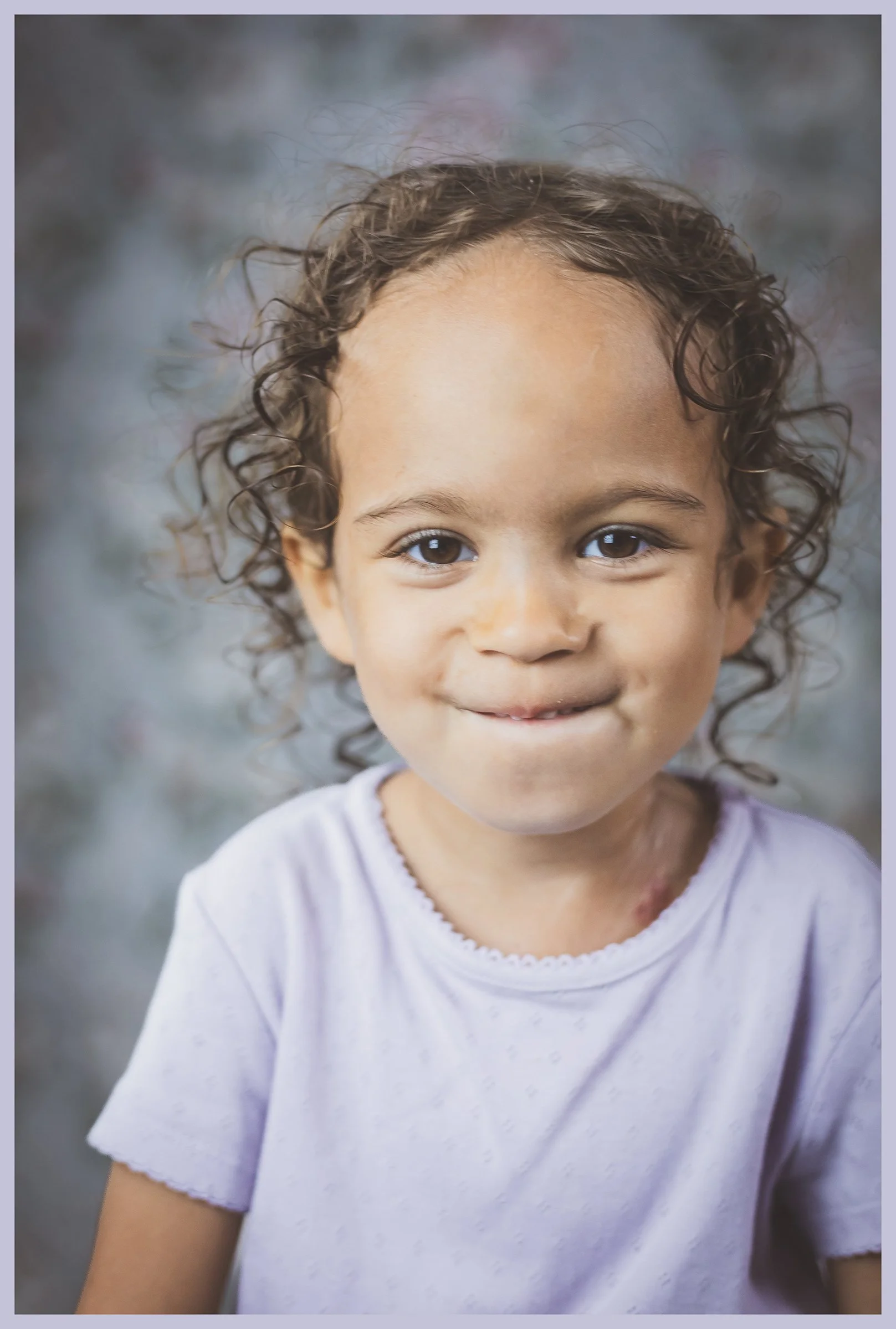 Close-up portrait of a young girl with curly hair and a gentle smile, wearing a white t-shirt, standing outdoors with a blurred natural background.
