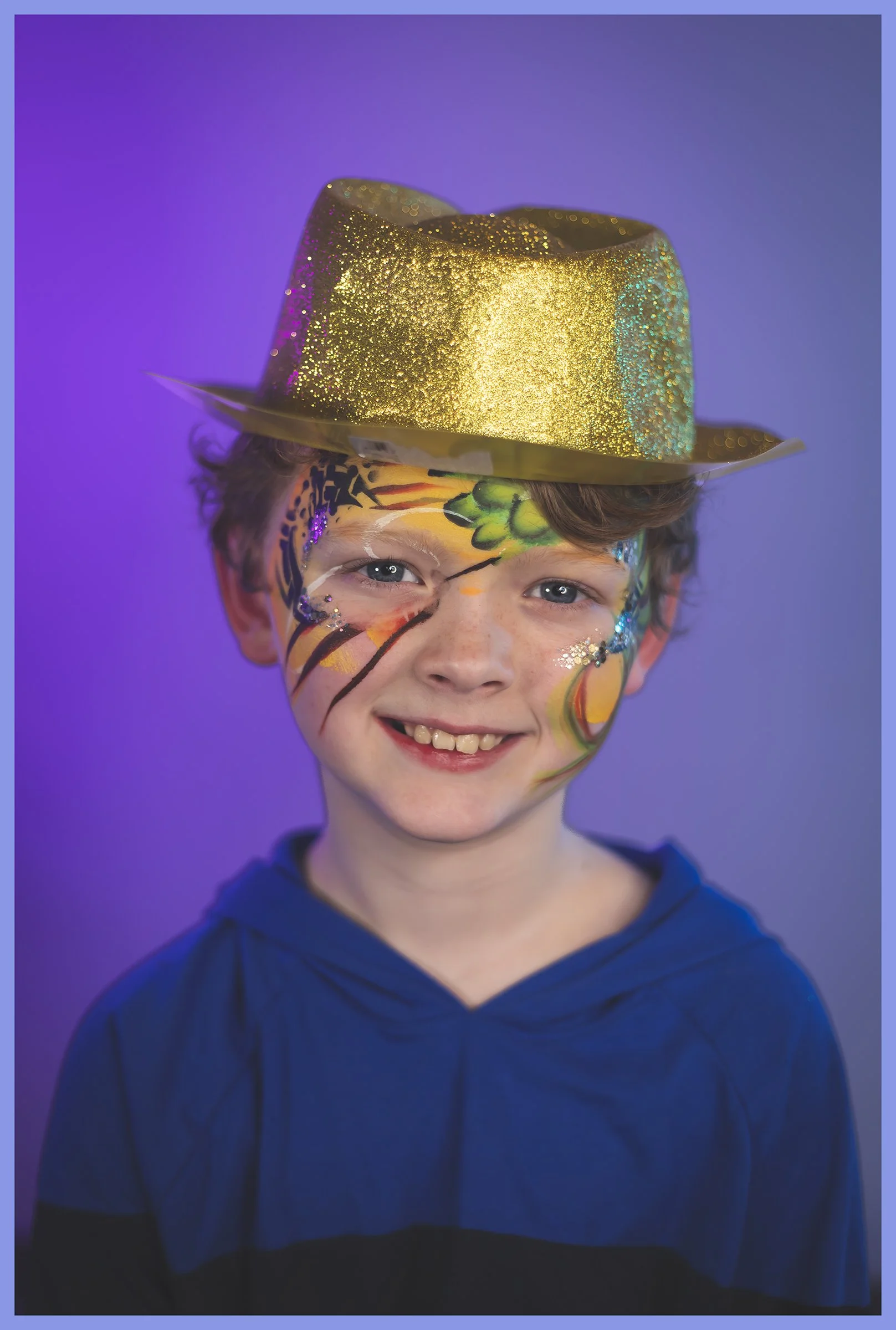 A young boy with colorful face paint and a big smile, wearing a shiny gold glittery hat, and a blue hoodie, against a purple background.