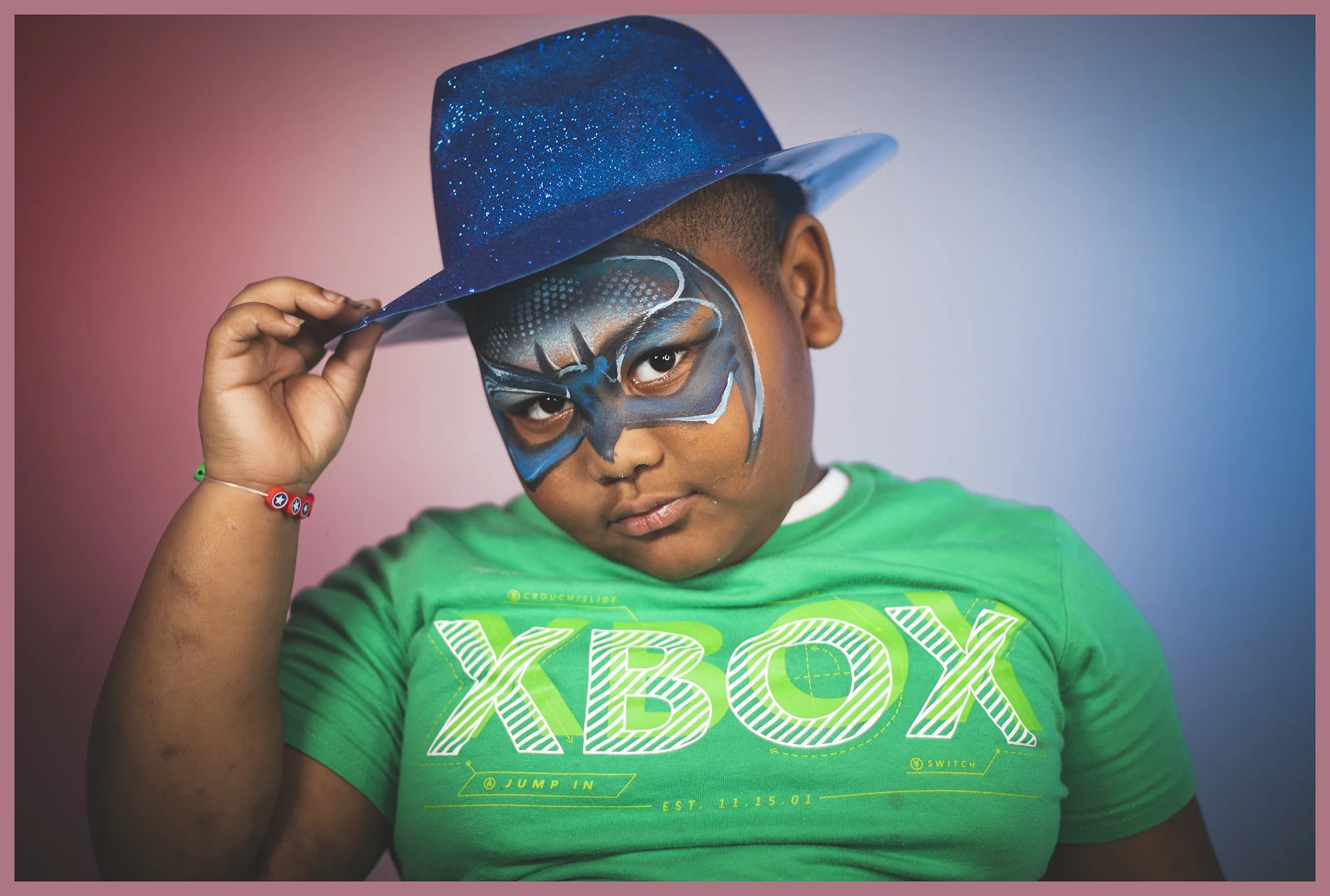 A young boy wearing a green XBOX t-shirt and a blue glittery hat, holding the hat with one hand. He has face paint resembling a superhero mask around his eyes, with a serious expression against a pink and blue background.