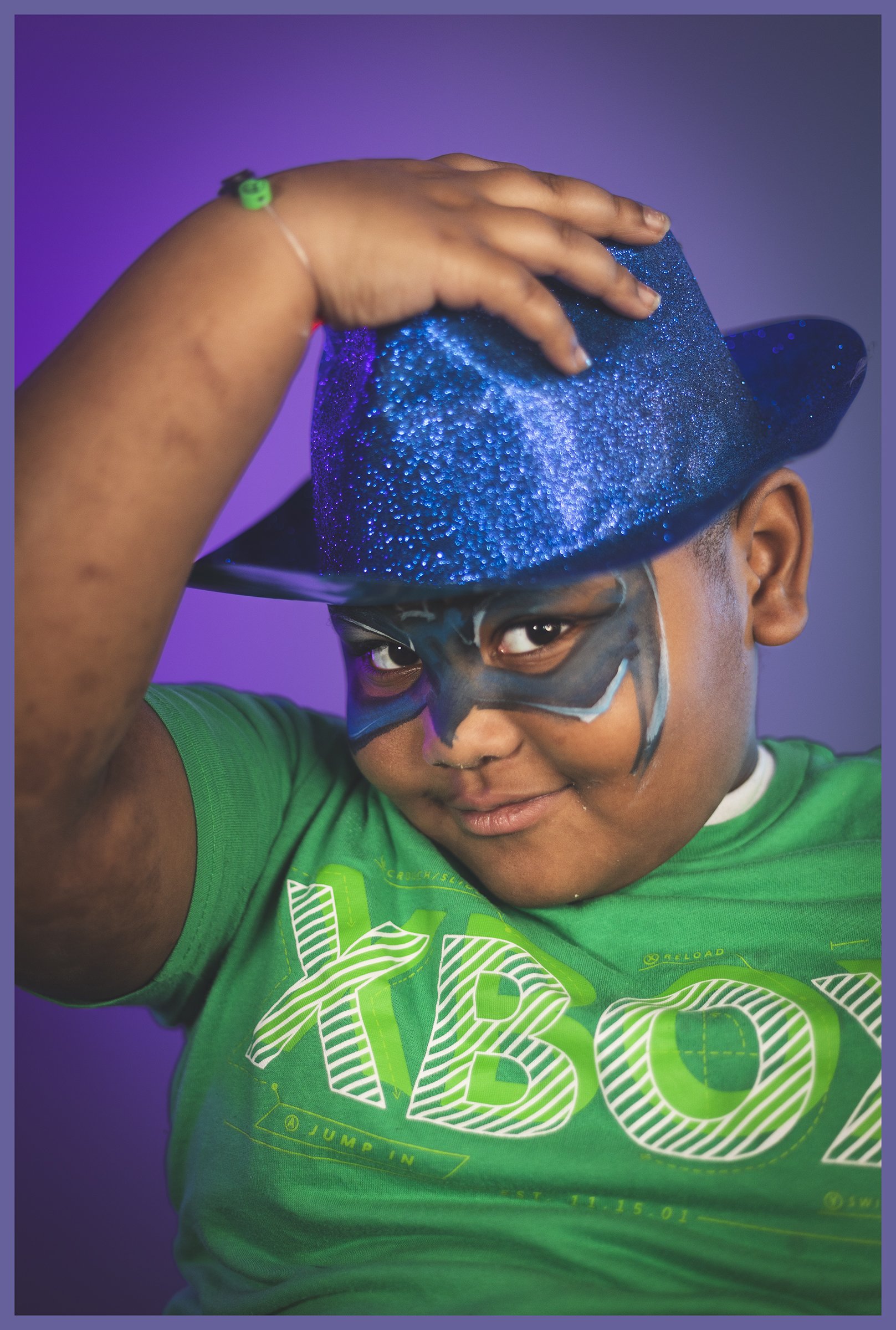 A young boy wearing a green T-shirt with a playful design, a glittery blue cowboy hat, and face paint resembling a superhero mask, posing with a confident expression.
