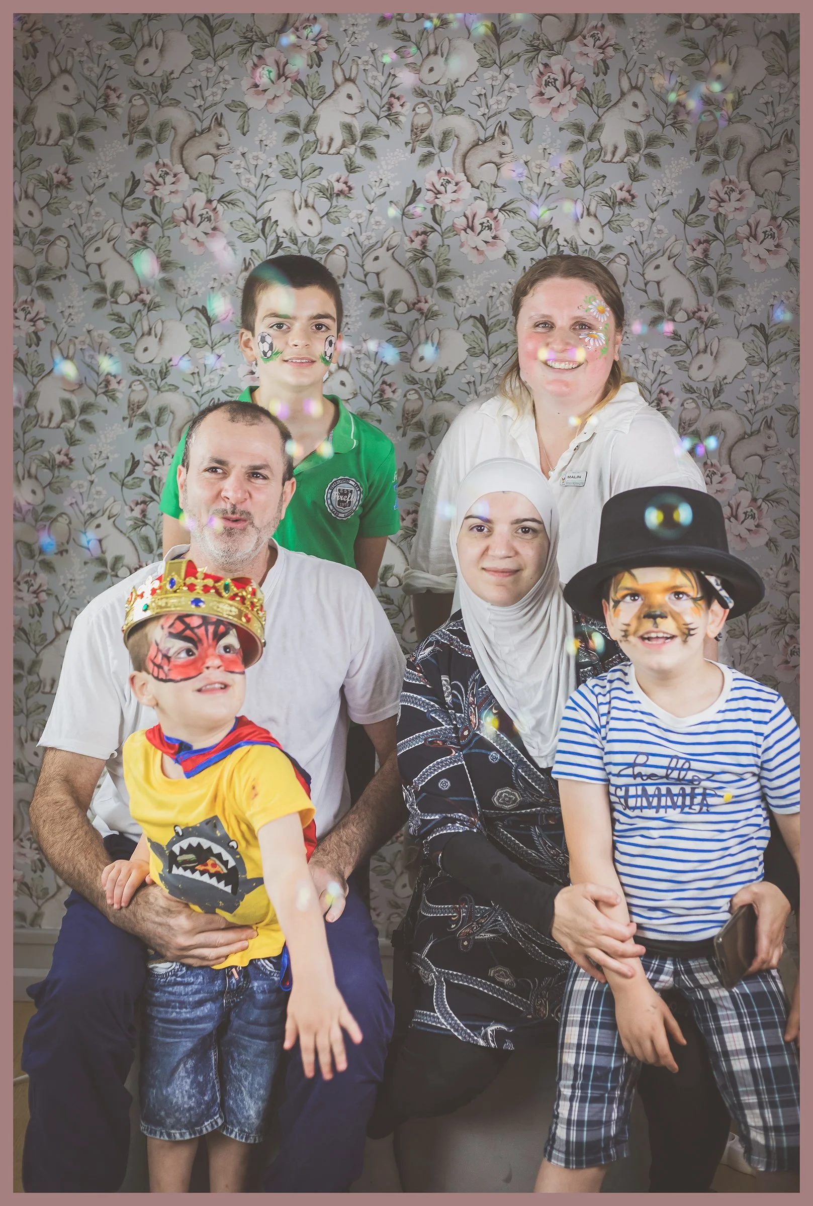 A group of seven people, including children and adults, celebrating at a party with fun face paint and costumes, standing in front of a bunny and floral wallpaper background, surrounded by bubbles.