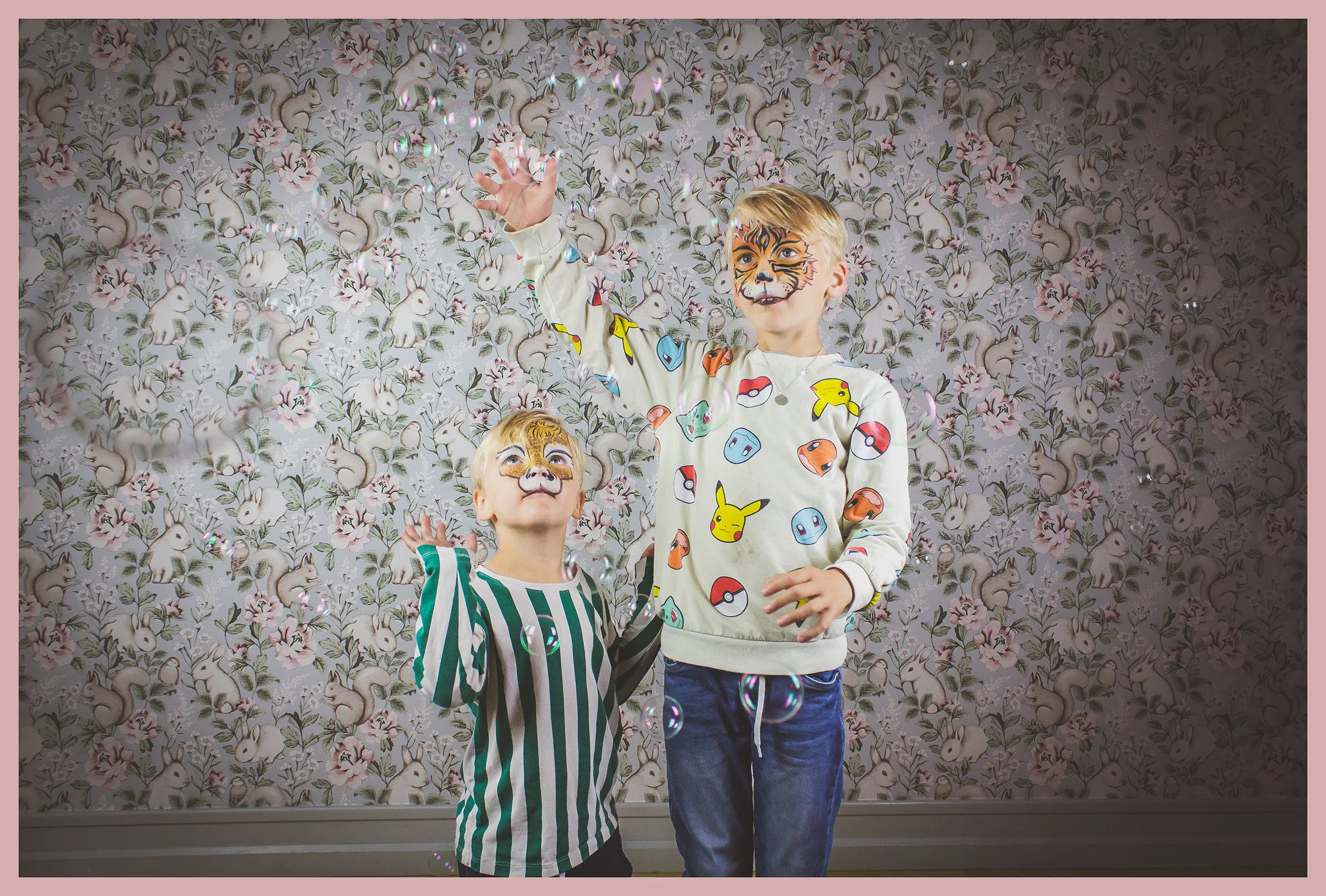 Two children with face paint of a tiger, playing with bubbles in front of a floral patterned wallpaper.