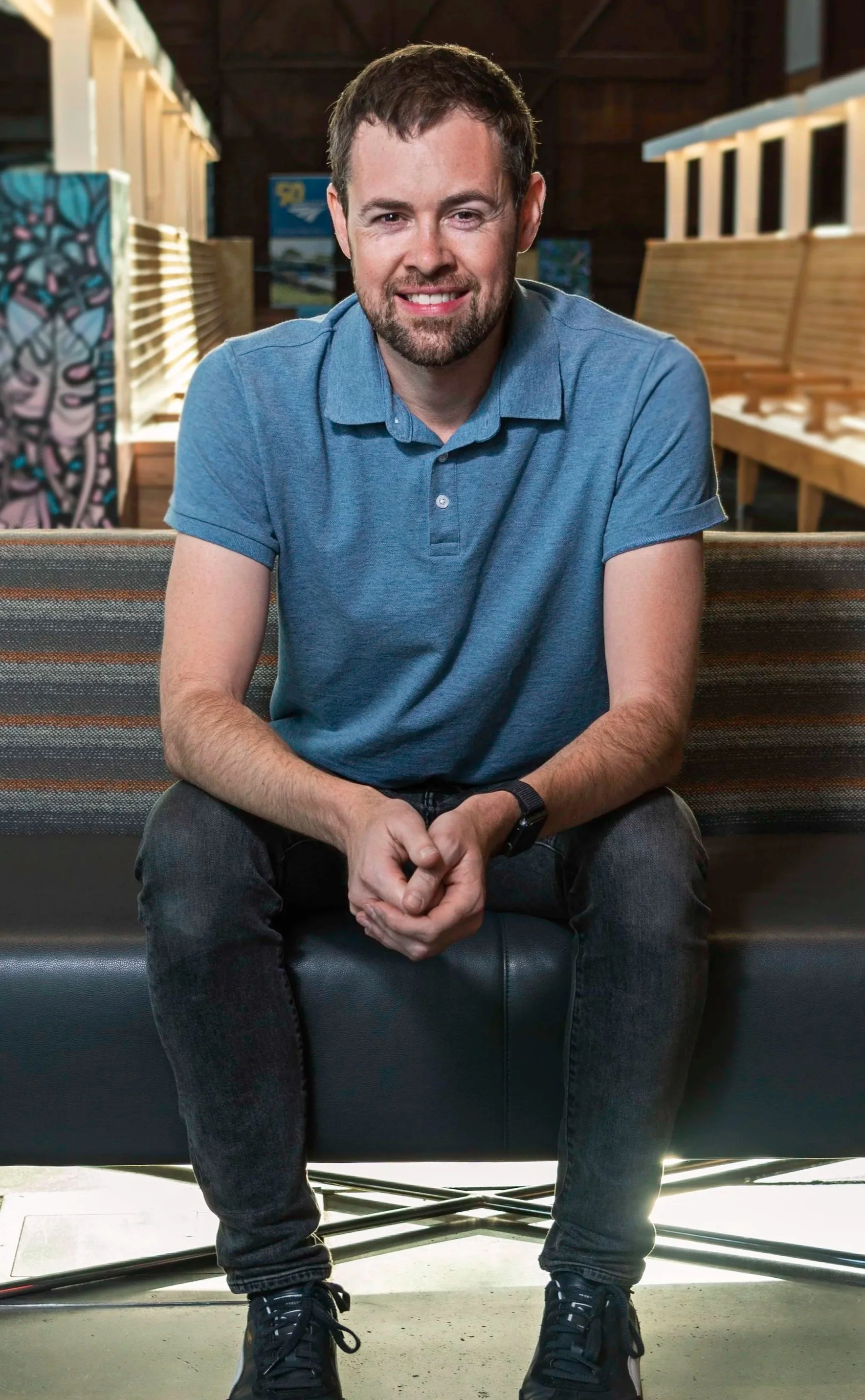 A man sitting on a bench inside a room, smiling, wearing a blue polo shirt, black jeans, and black shoes, with artwork and wooden benches in the background.