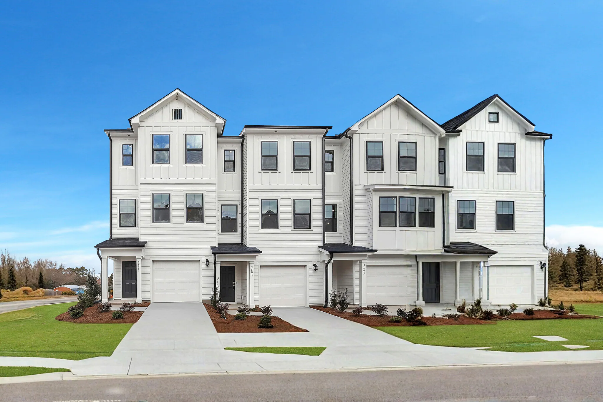 White multi-story townhouses with black roofs and black doors, front lawns with small bushes, concrete driveways, and a blue sky.