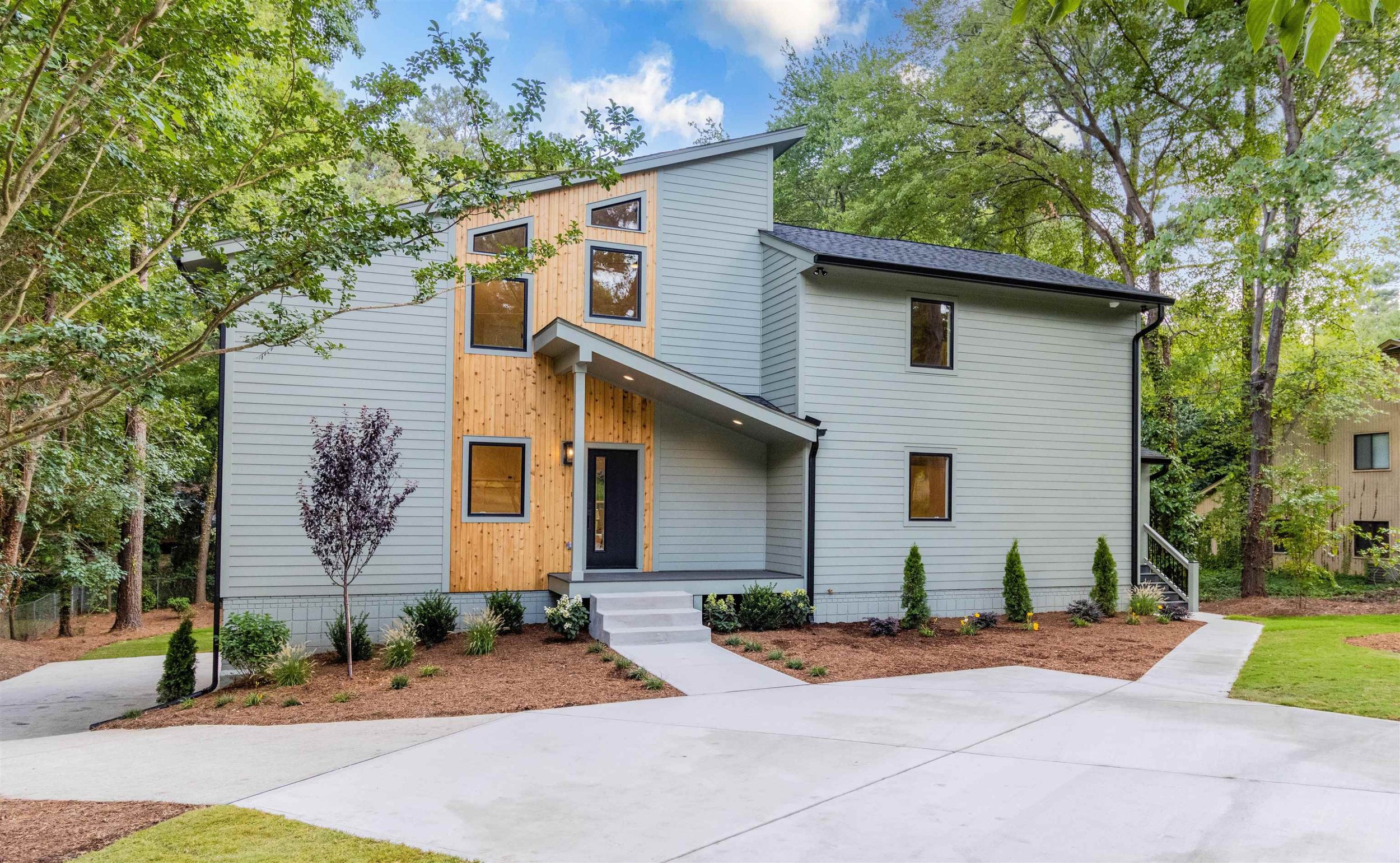 Modern two-story house with gray siding and wooden accents, surrounded by landscaped yard with trees and bushes.