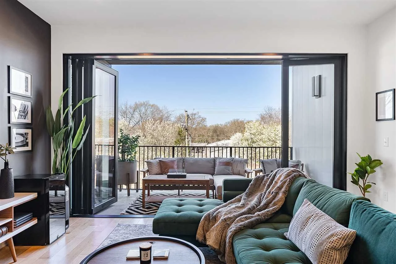 Living room with an open sliding glass door leading to a balcony with outdoor seating and trees in the background.