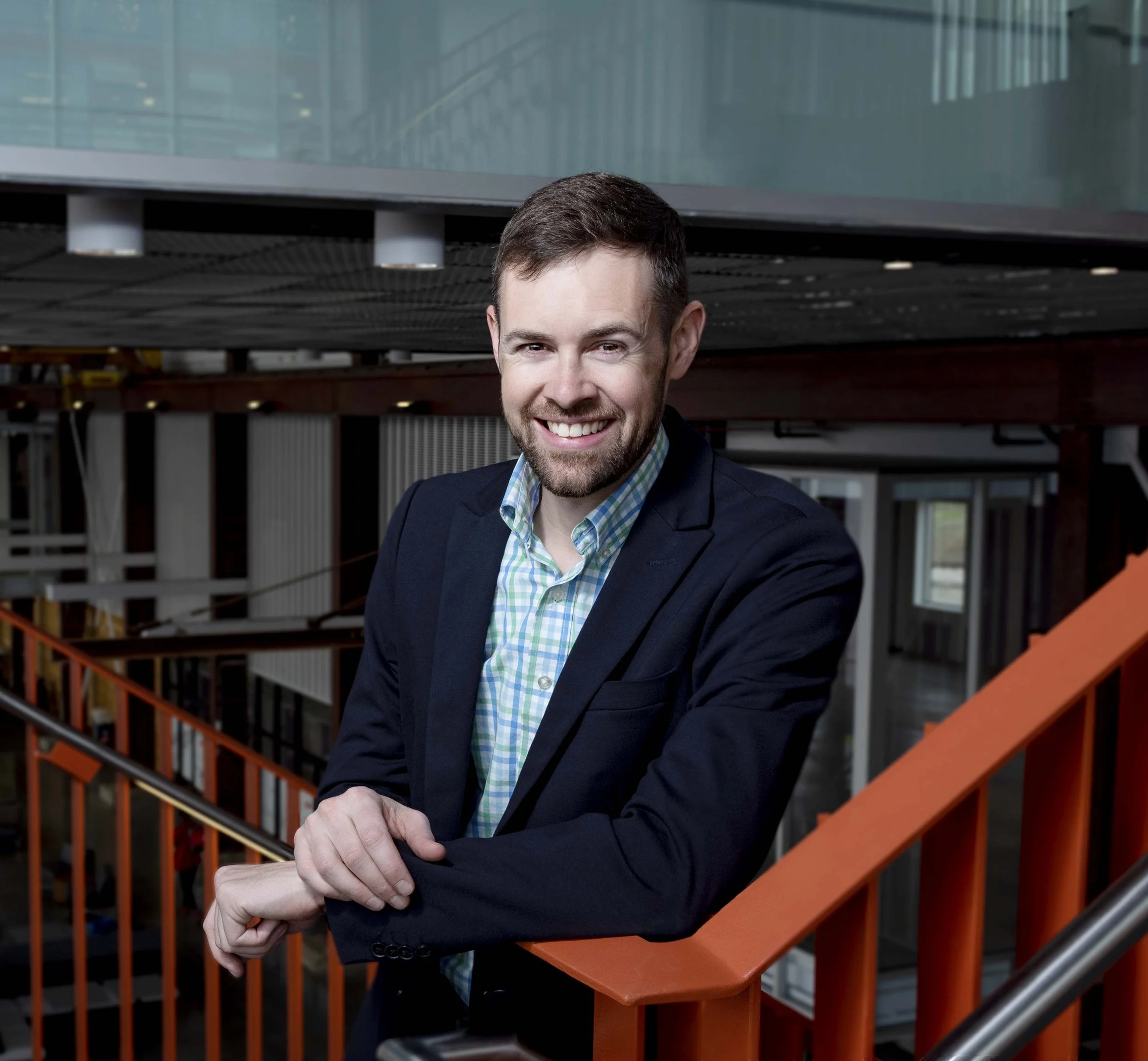 A young man with brown hair and a beard smiling, wearing a dark blazer over a light green and blue checkered shirt, standing on an orange staircase inside a modern office building.