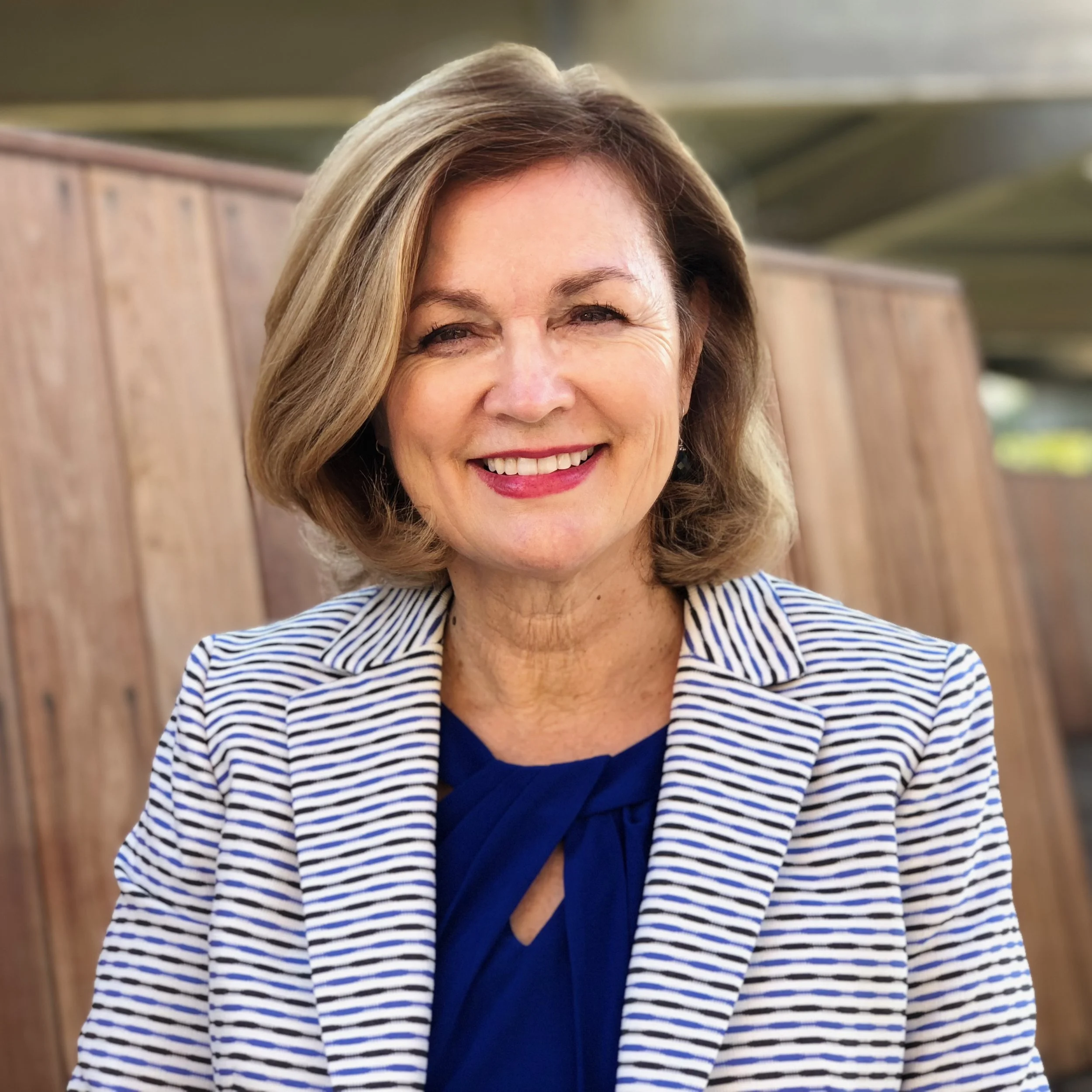A smiling middle-aged woman with shoulder-length blonde hair, wearing a blue top and a white and navy striped blazer, standing outdoors with a wooden fence in the background.