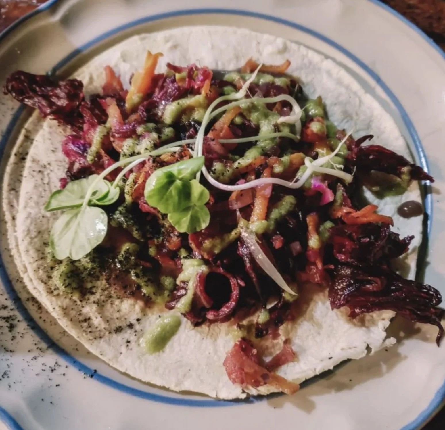 A close-up of a taco topped with shredded meat, diced vegetables, microgreens, and a drizzle of green sauce, served on a white and blue plate.