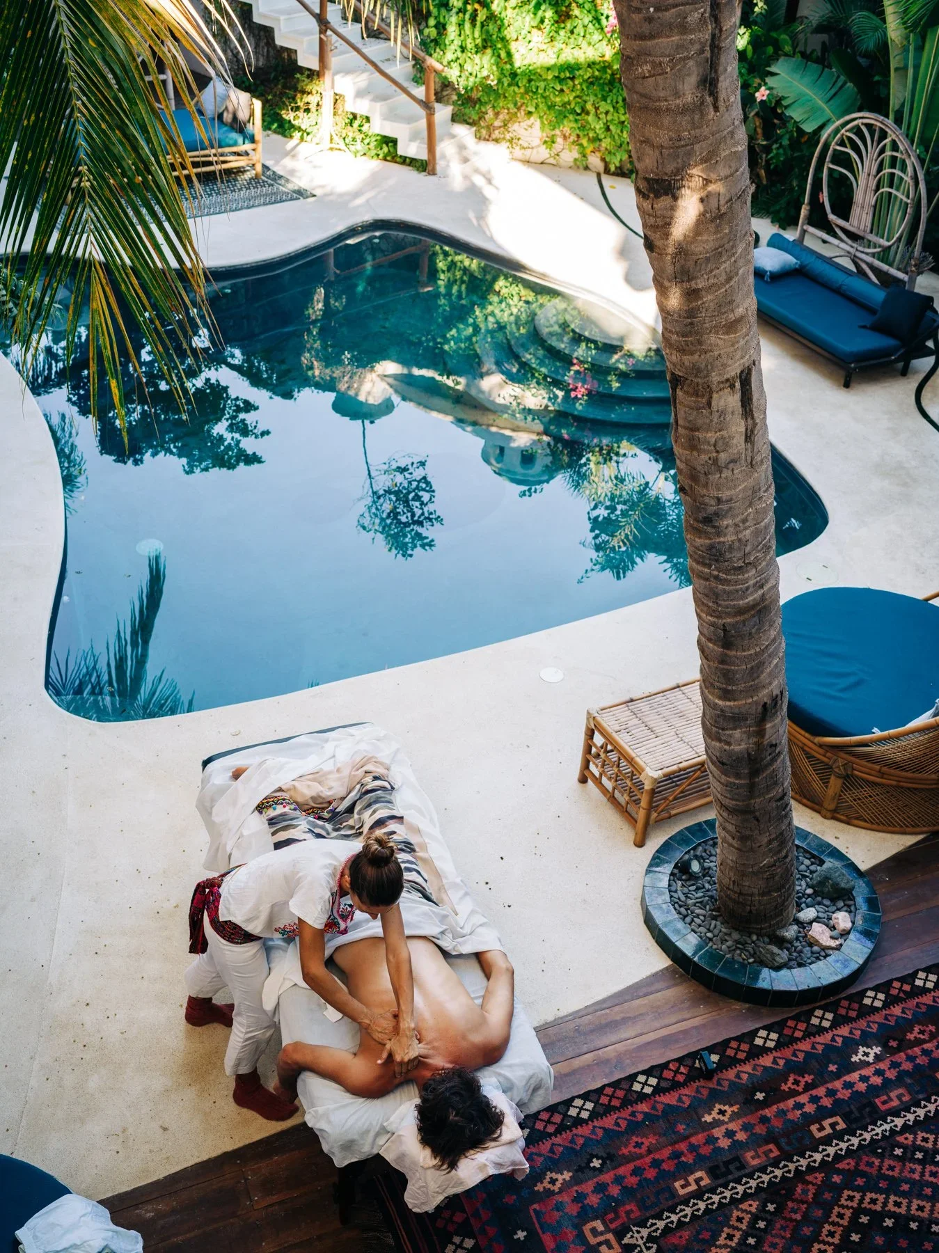 A woman receiving a massage outdoors beside a swimming pool, with patio furniture, a tree, and lush greenery in the background.