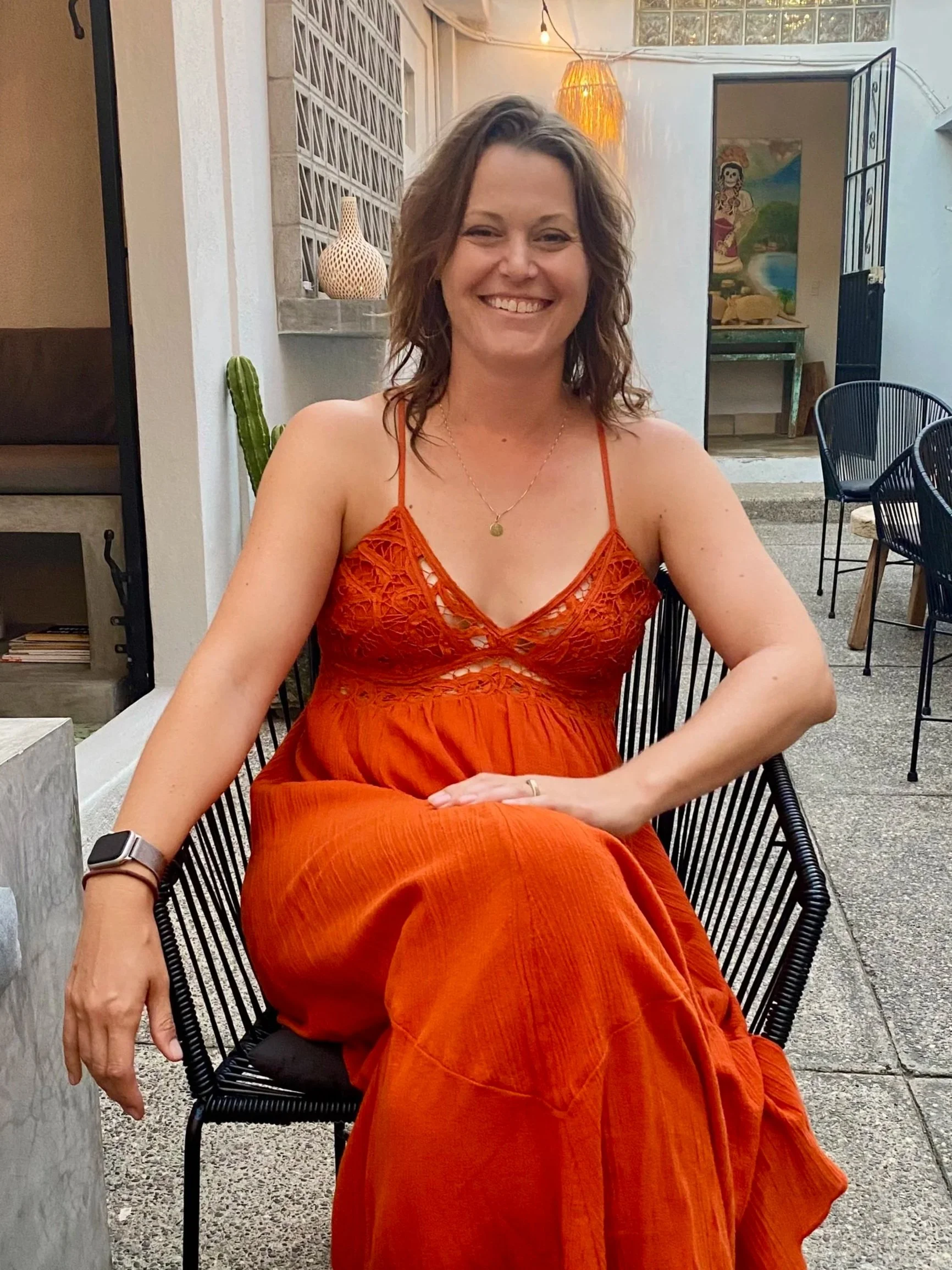 A woman with shoulder-length curly hair wearing a bright orange dress with lace detail, sitting outdoors on a black chair with a relaxed posture, smiling at the camera.