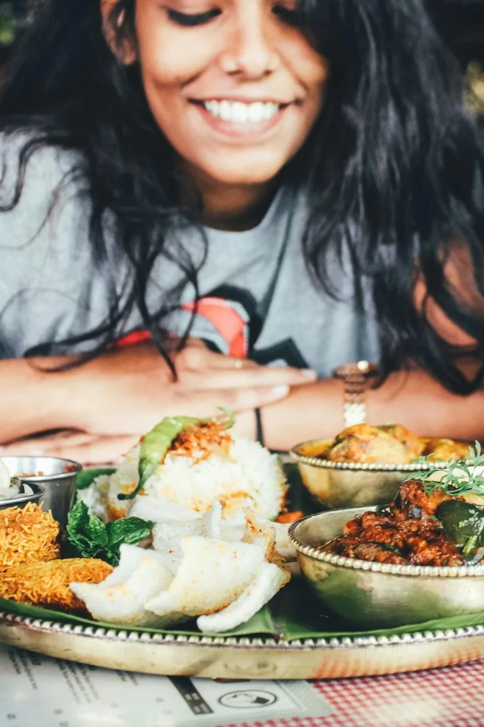 A woman with long black hair smiling at a table with Indian dishes, including rice, curry, and naan bread.