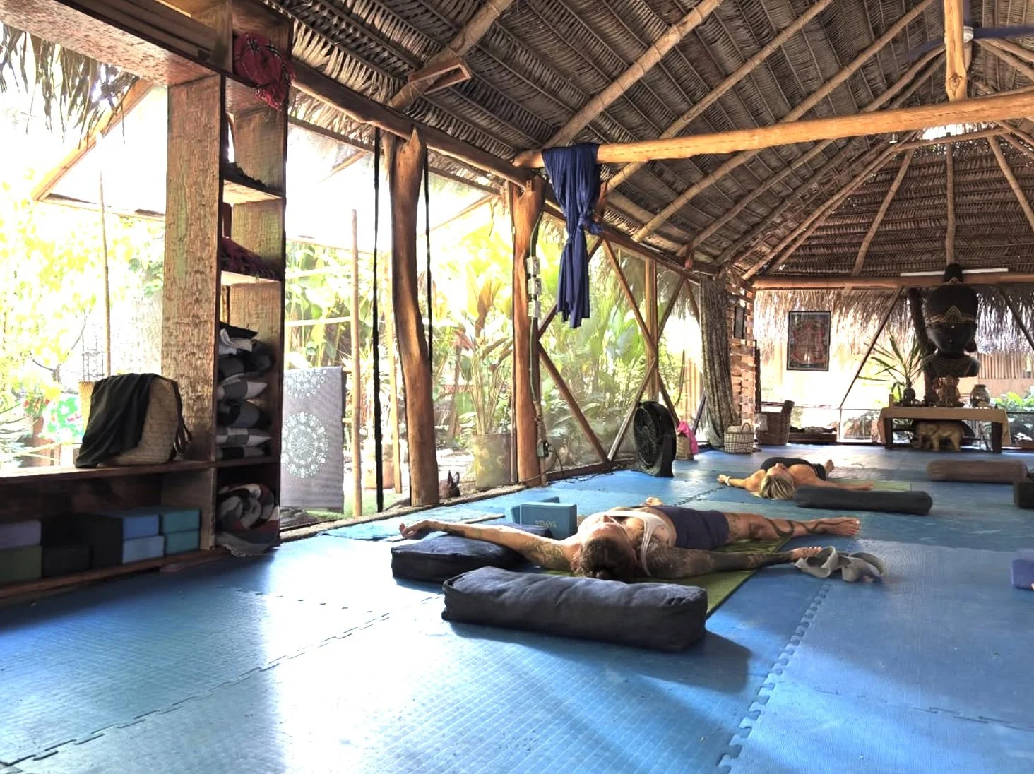 People practicing yoga in a bright, open-air studio with a thatched roof, wooden beams, and large windows showing lush greenery outside.