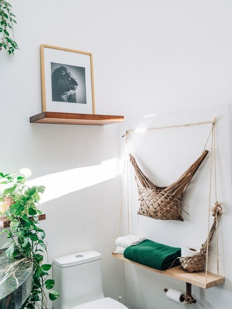A minimalist bathroom with a white wall, a framed black-and-white lion photograph on a floating wooden shelf, a potted plant on a marble countertop, a toilet, and a wooden swing seat with a hammock-style basket hanging on the wall, with a green towel on the seat.