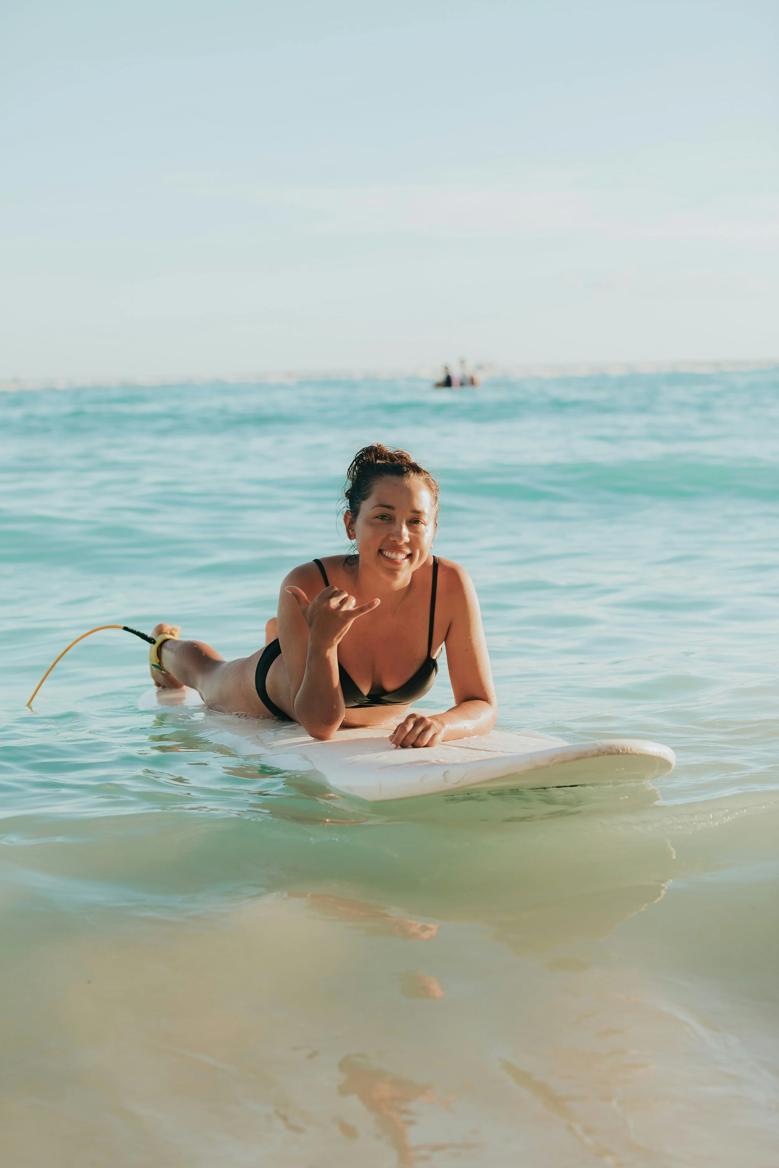 A woman lying on a surfboard in the ocean, smiling and making a call me hand gesture with her right hand while in black swimwear.