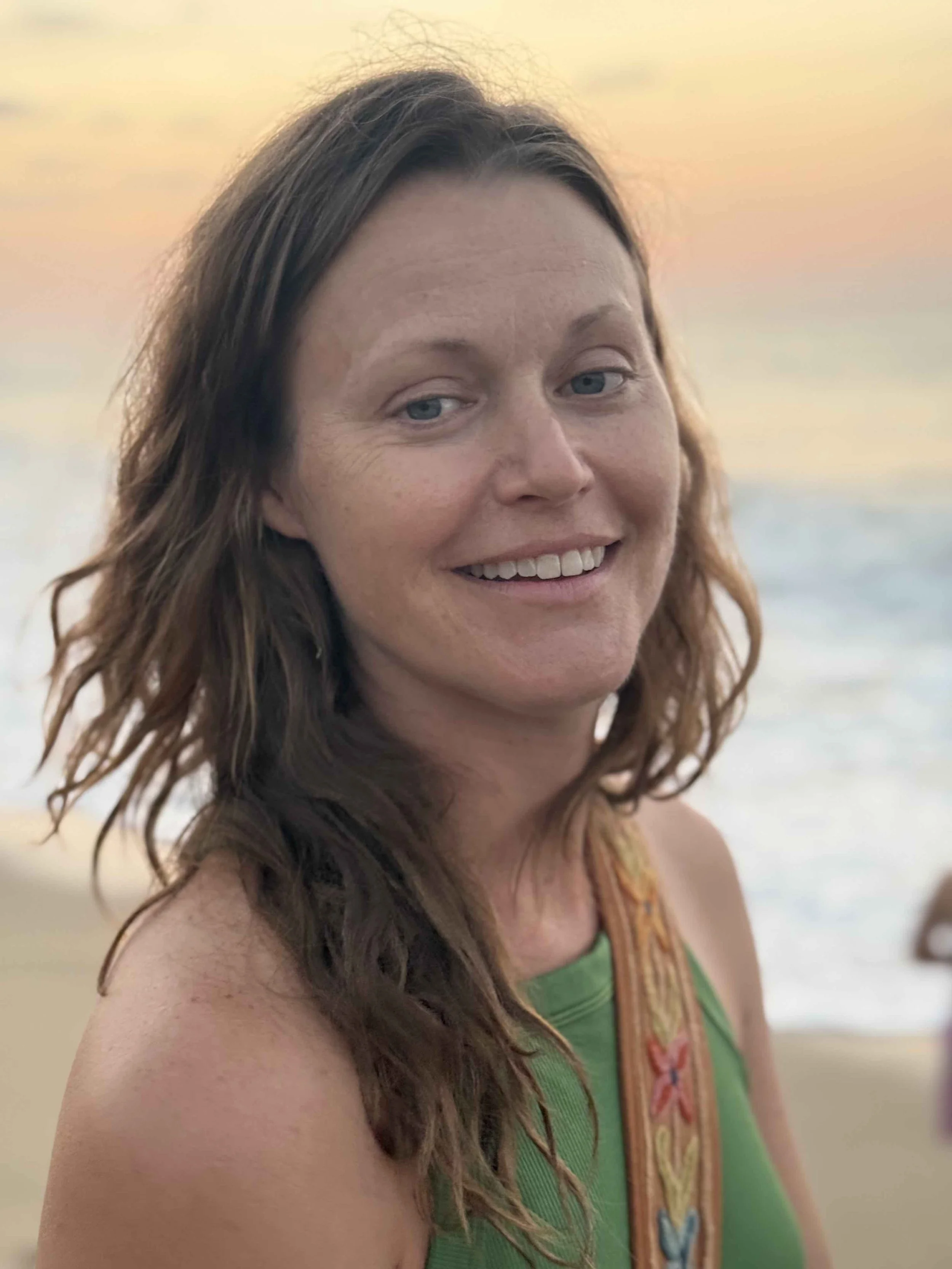 A woman with wavy brown hair and blue eyes smiling at the camera at the beach during sunset.