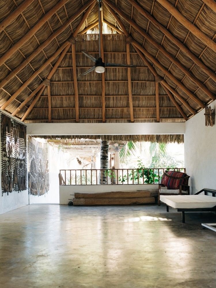 An open-air room with a thatched roof, ceiling fan, and wooden flooring. There is a balcony with a railing overlooking lush greenery outside, decorated with textiles and wooden furniture.