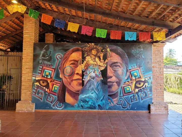 Colorful mural featuring two large human faces, a woman dressed in traditional indigenous clothing, and various geometric and cultural symbols. Decorations include colorful paper banners hanging from the ceiling.