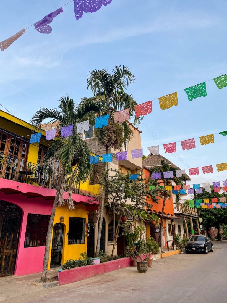 Colorful residential buildings with pink, yellow, and orange facades, decorated with colorful hanging papel picado flags, palm trees, and potted plants on a street under a blue sky.