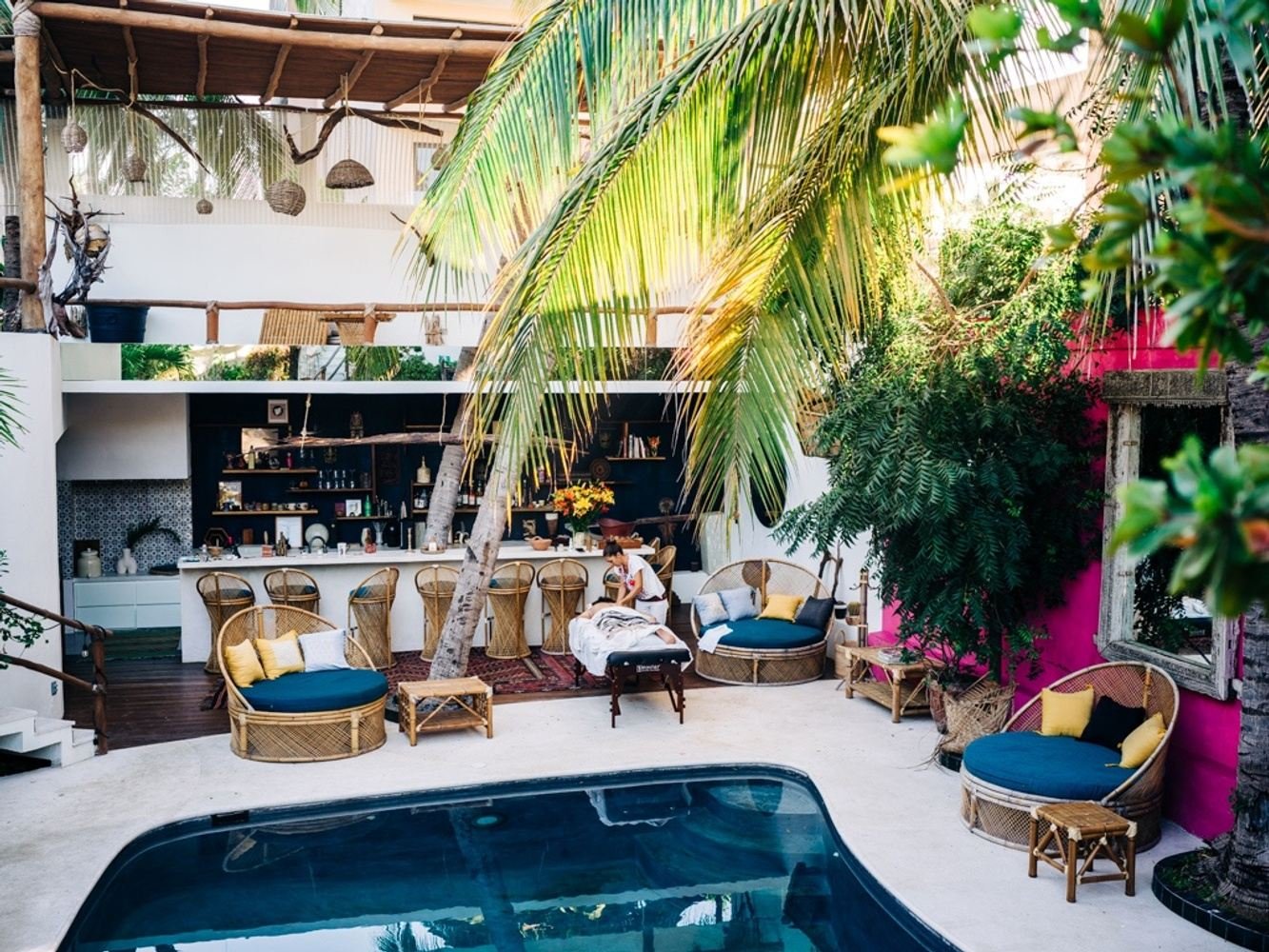 Outdoor tropical poolside lounge area with wicker furniture, blue cushions, yellow pillows, bar with stools, lush green palm trees, and a pink wall.