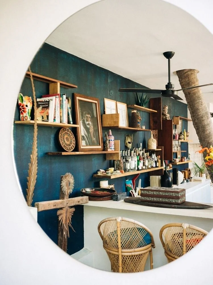 Interior view of a room seen through a circular opening, featuring a blue accent wall with wooden shelves holding books, framed artwork, and decorative items, a bar with various bottles, and wicker chairs.