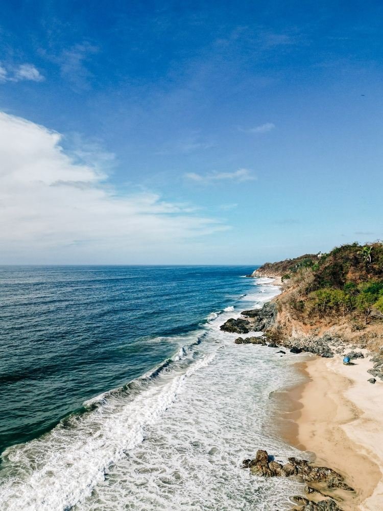 Coastline with sandy beach, rocks, and cliffs under a partly cloudy blue sky