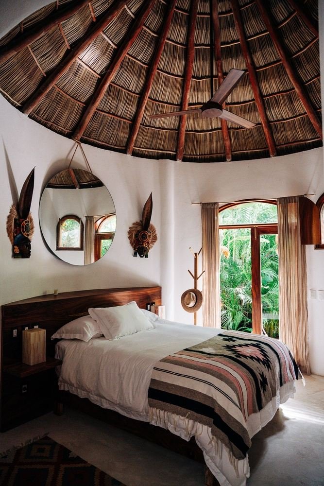 A cozy bedroom with a thatched, circular ceiling, a bed with white bedding and a tribal patterned blanket, a wooden headboard, side tables, a mirror, and large windows with beige curtains showing lush greenery outside.