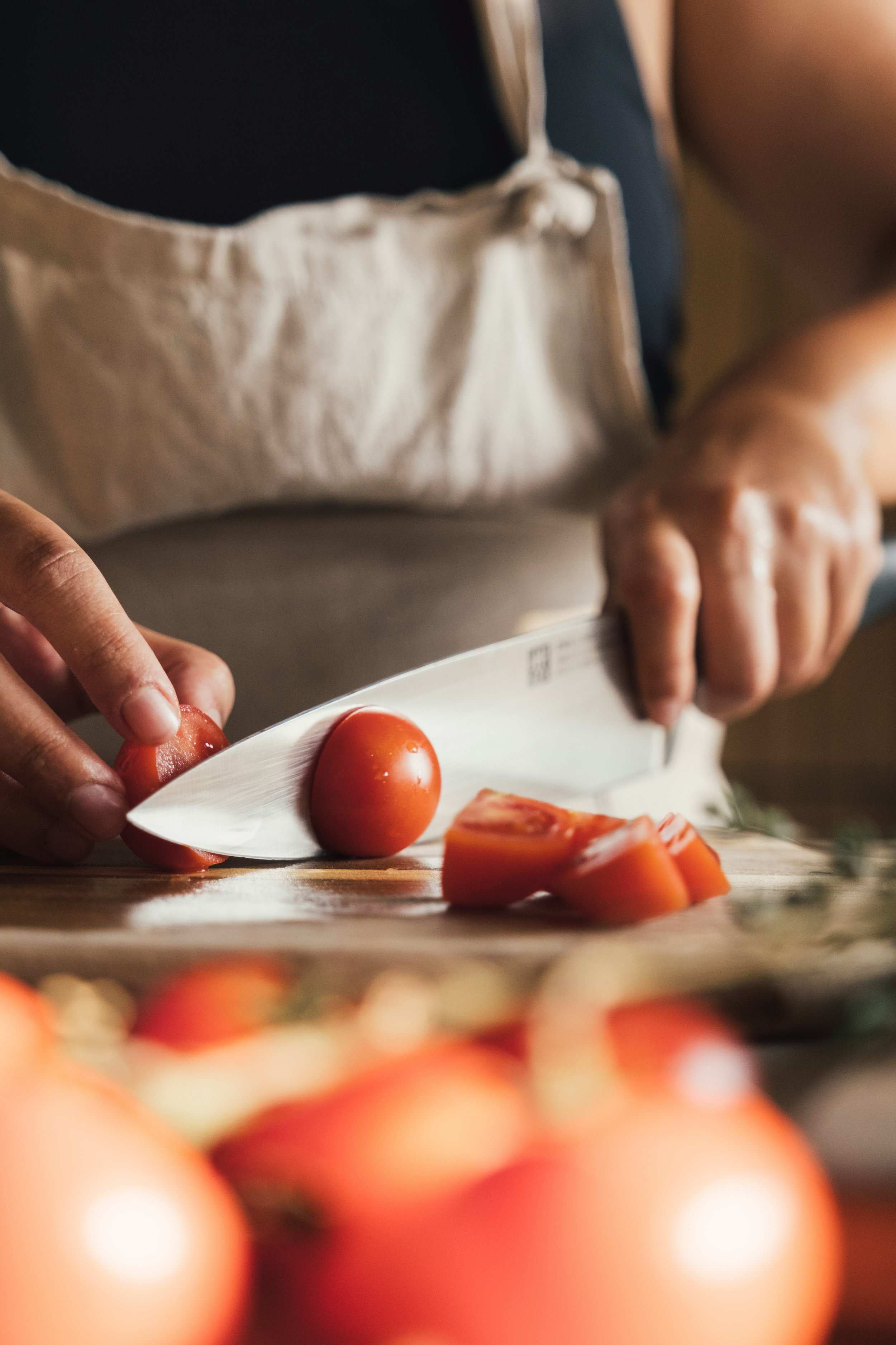Person slicing a cherry tomato on a cutting board with a chef's knife, with whole cherry tomatoes and other vegetables in the foreground.