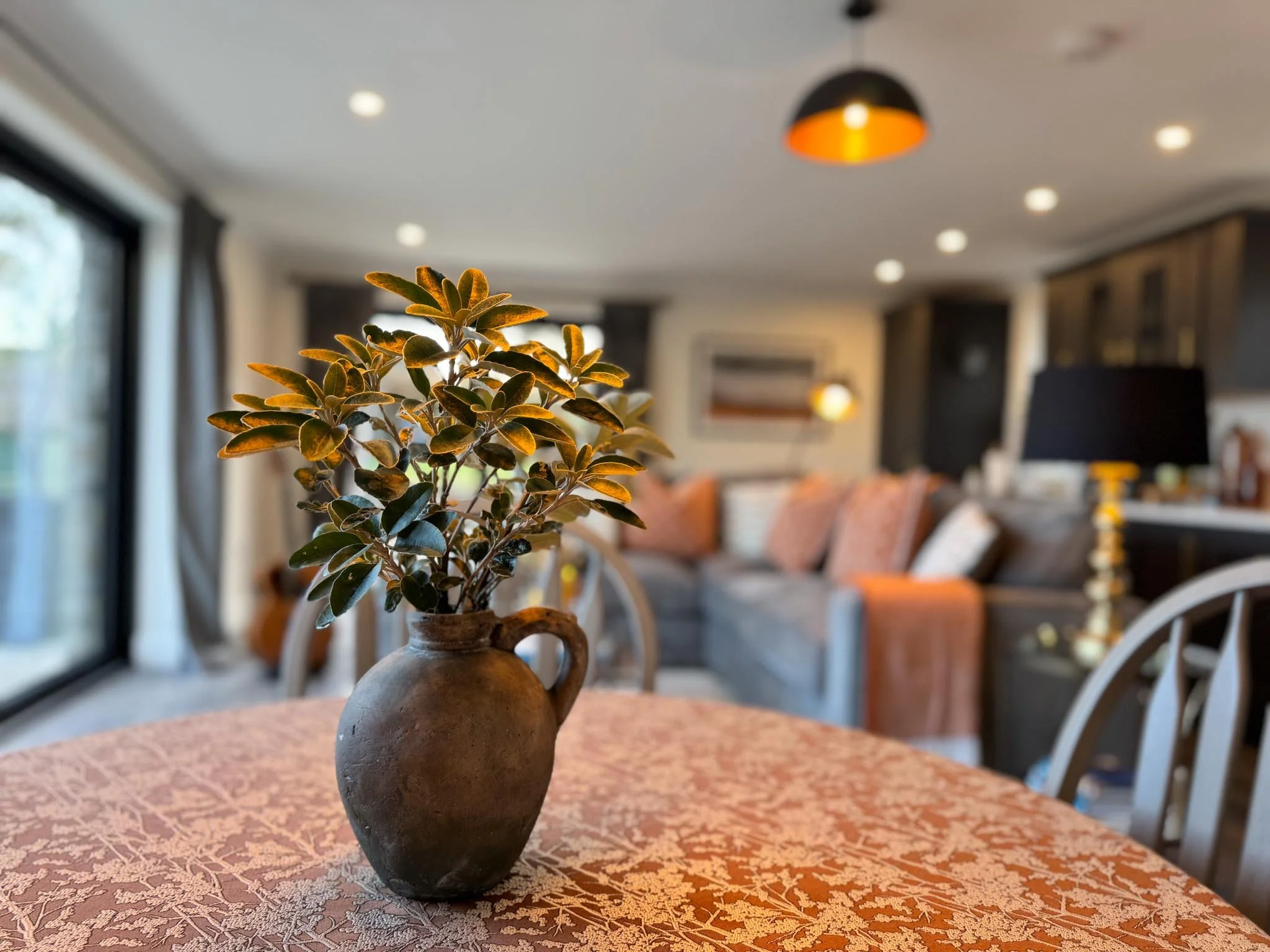 A brown ceramic vase with green leafy plant on an orange patterned table in a living room with pillows, lights, and window in the background.