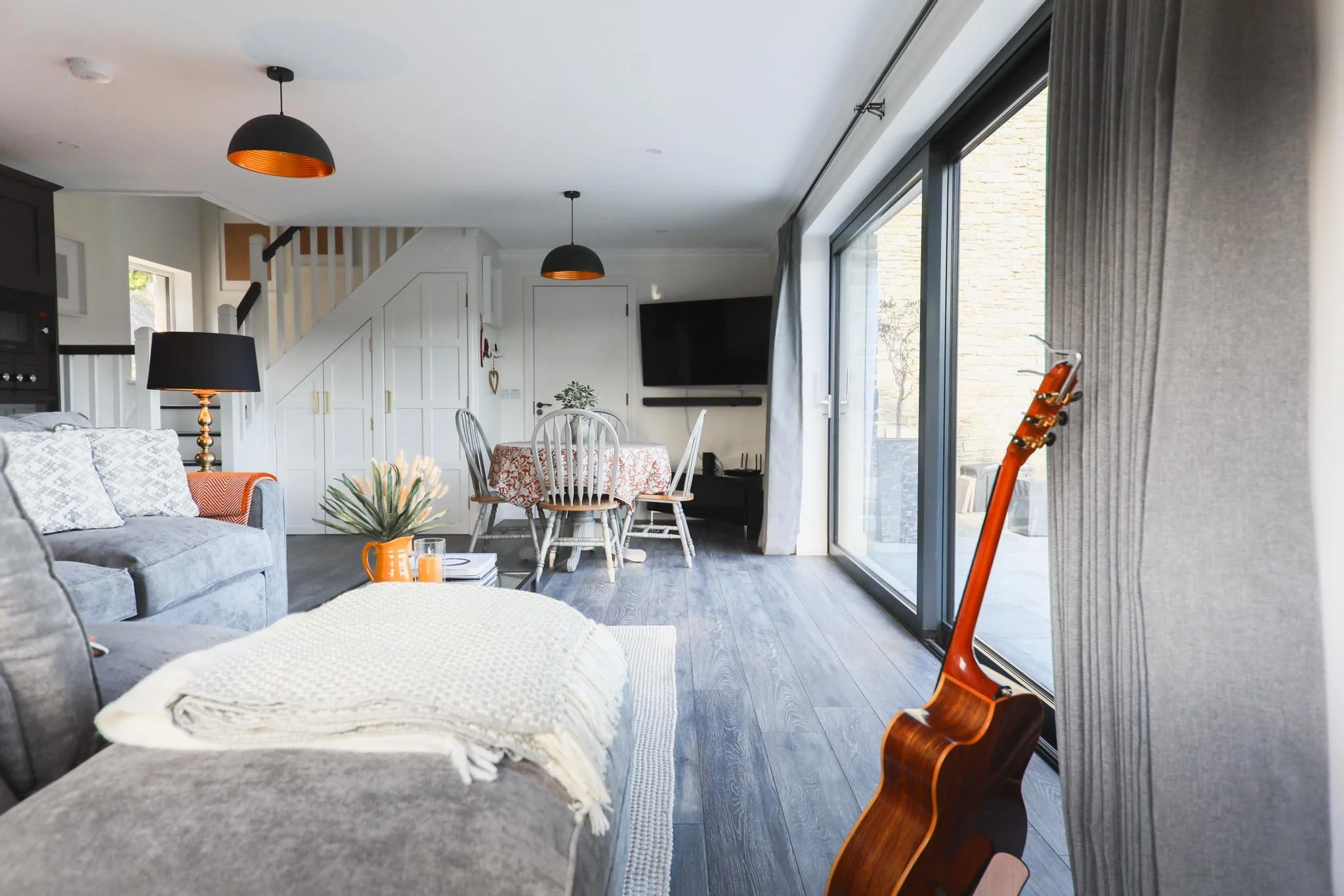 Modern living room with large glass sliding doors, gray sofa with throw blankets, a guitar on the floor, a coffee table with an orange pitcher and books, a dining table with six chairs, and a black radio and TV on the wall, in a bright, airy space.