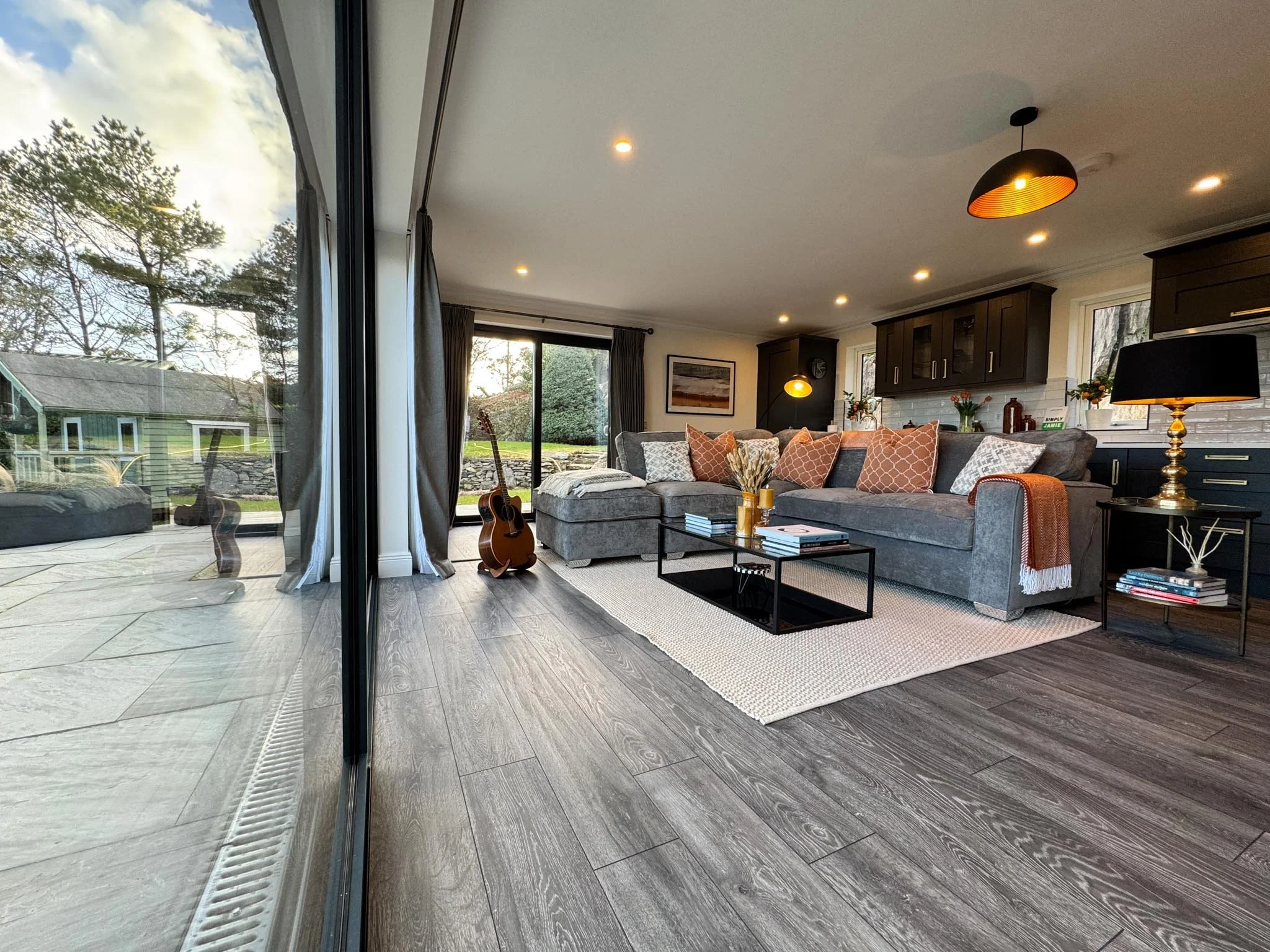 Living room with large window, gray sectional sofa with multiple pillows, coffee table, guitar, sliding glass door leading outside, modern kitchen in the background, hardwood floors, various lighting fixtures, and decorative accents.