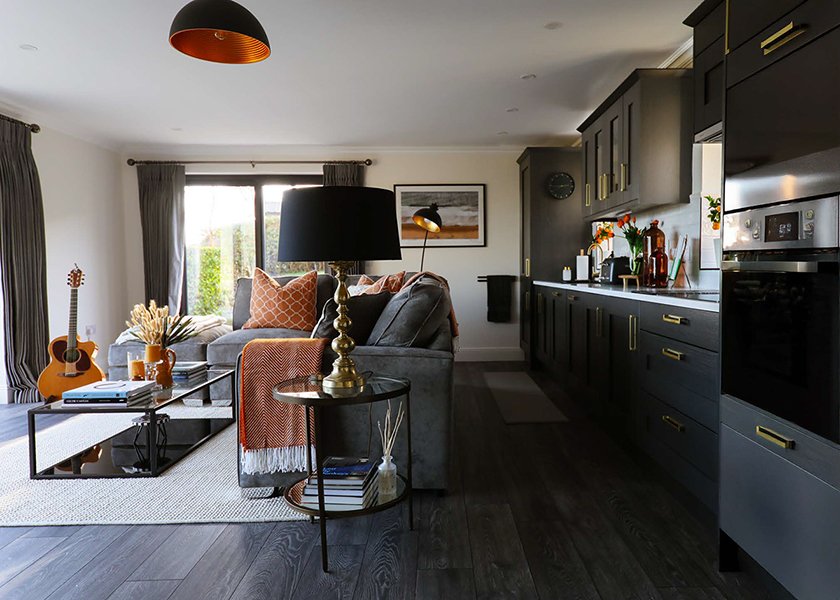 Living room with gray sofa, black coffee table, guitar, and orange pillows, open to a kitchen with dark cabinets, countertop decor, and a window with curtains