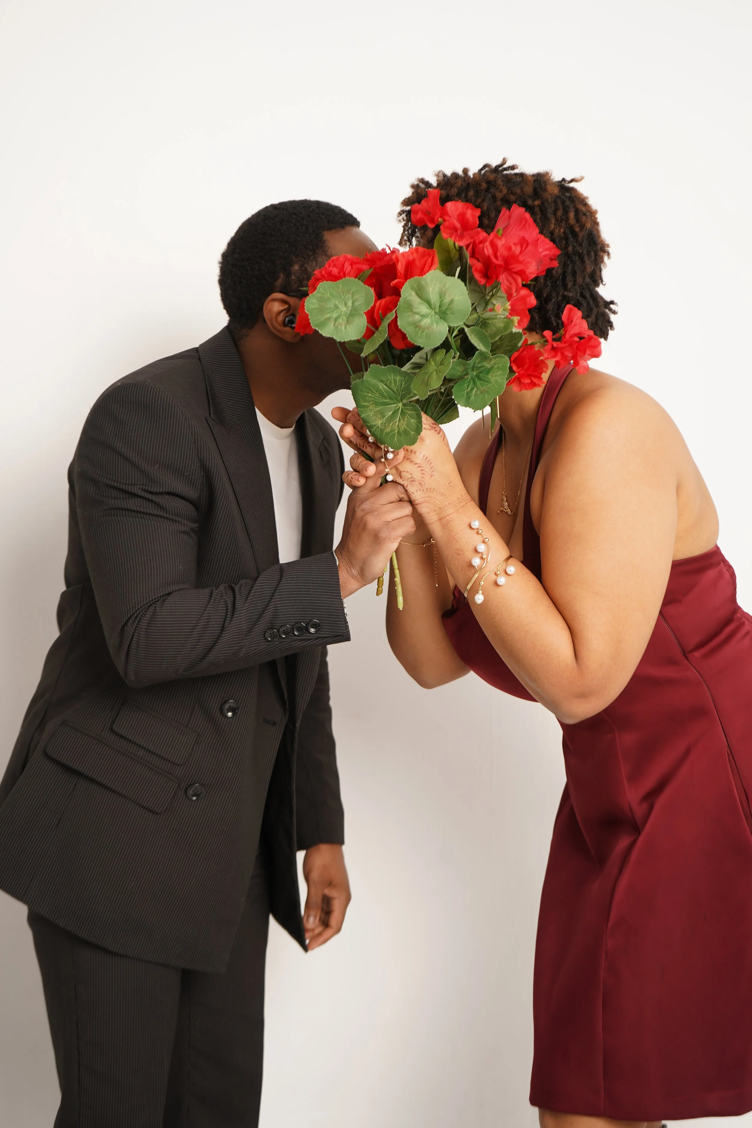 A man and a woman share a kiss, with the woman's face obscured by a bouquet of red flowers they are holding between them, against a plain white background.