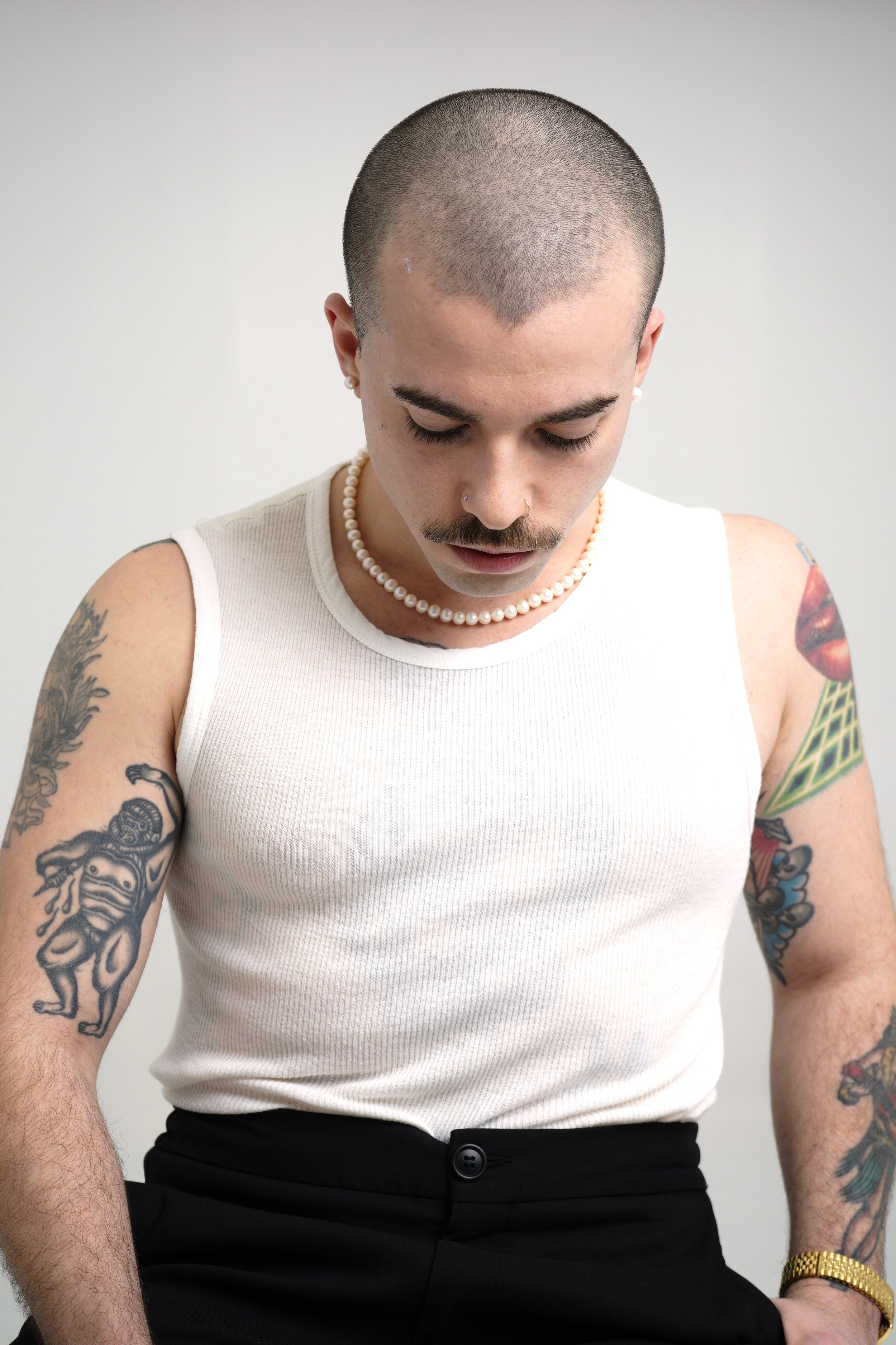 A person with a buzz cut hairstyle, tattoos on both arms, wearing a white sleeveless shirt, pearl necklace, and gold watch, with their head bowed down.