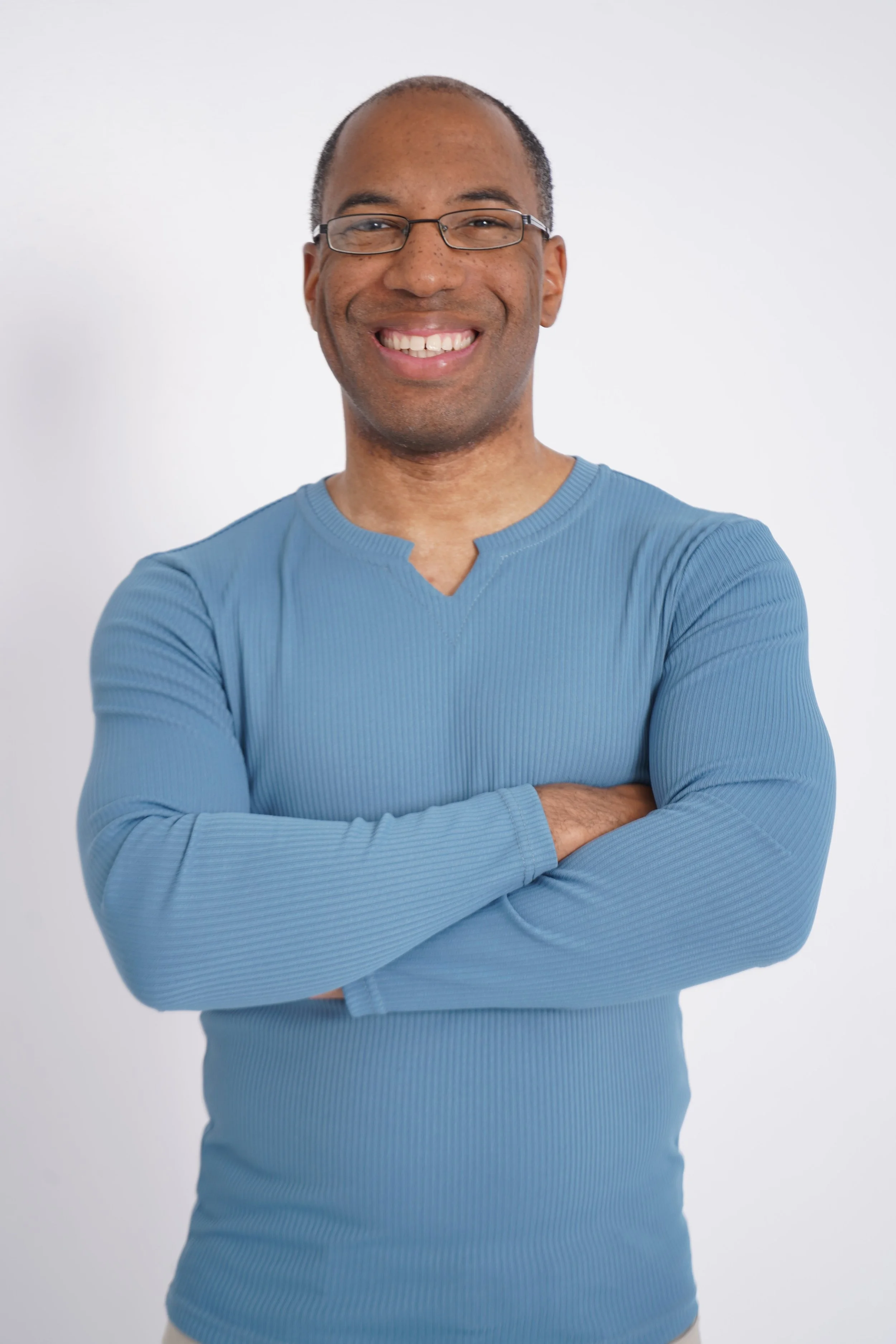 Smiling man wearing glasses and a light blue, long-sleeve shirt with arms crossed in front of a plain white background.