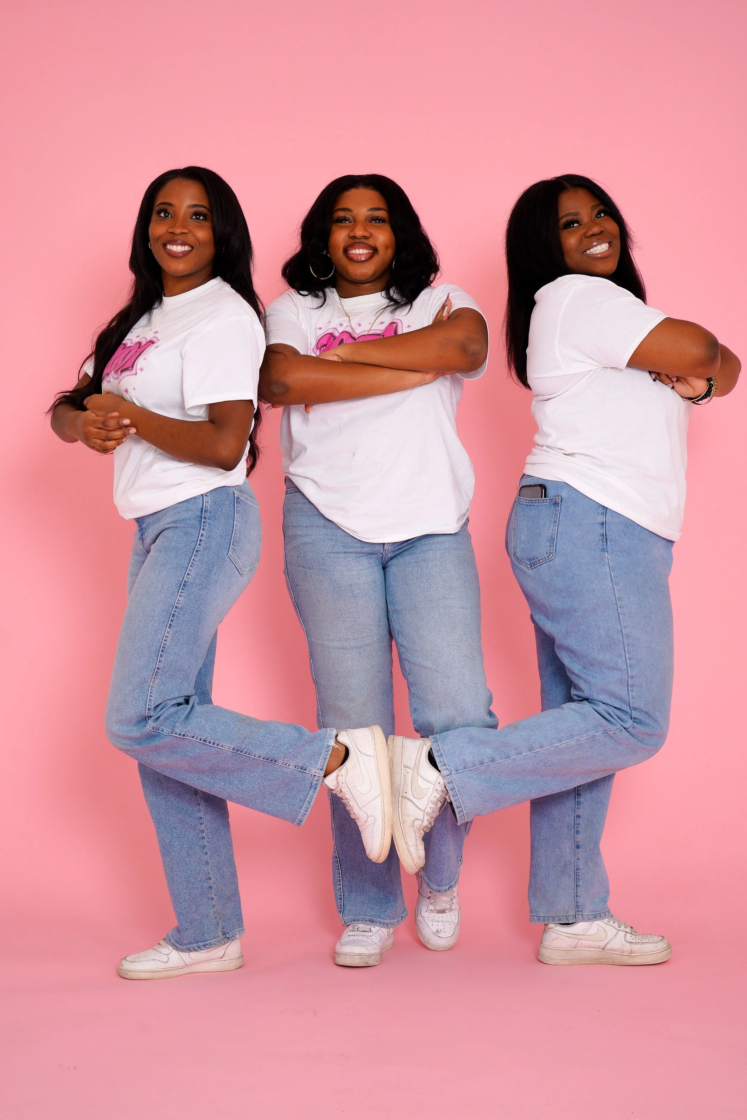 Three women in white T-shirts with pink graphics and light blue jeans, standing against a pink background. The woman in the middle has her arms crossed, and the women on the sides are standing with one leg lifted, their feet touching the others' ankles. All are smiling.