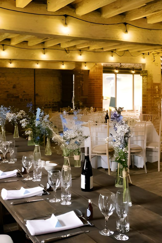 Interior of the Courtyard Barns Venue in Leicester, decorated for a special occasion, such as a wedding, with floral arrangements, wine bottles, glassware, and set tables under warm string lighting.