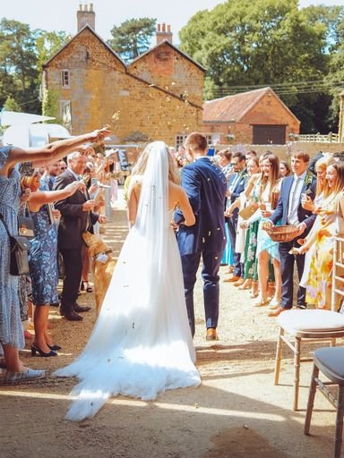 Wedding ceremony taking place at the Courtyard Barns Venue in Leicester with a bride and groom walking down an aisle lined with guests under clear skies, with old buildings and trees in the background.