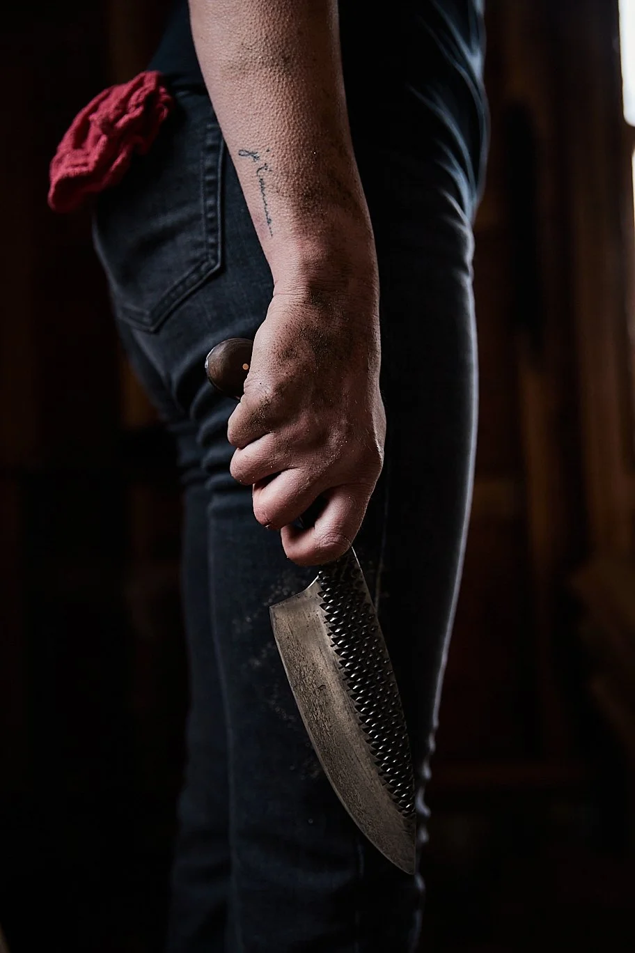 A person wearing black jeans holding a large, rusted meat cleaver in their right hand, with a red cloth tucked into the back pocket of their jeans.