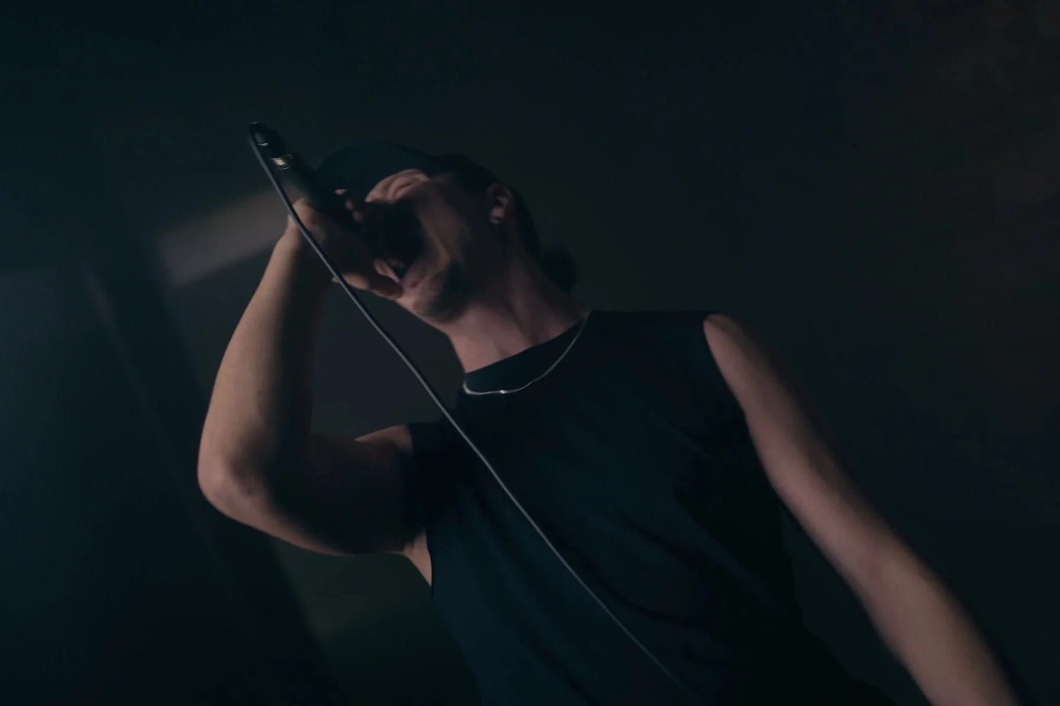 Ein Mann trägt eine schwarze Weste, einen schwarzen Caps und eine Halskette, er hält ein Mikrofon vor den Mund und singt in einem dunklen Raum.