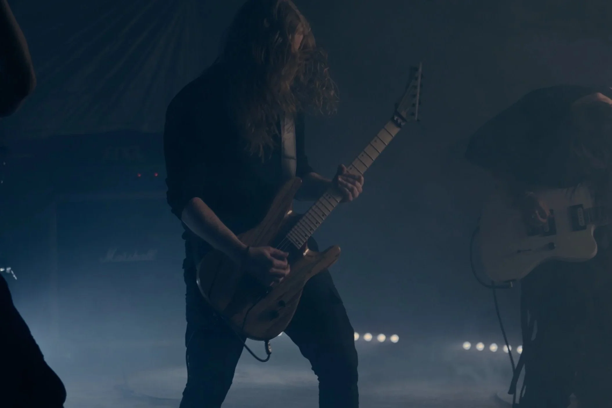 Ein Musiker mit langen Haaren spielt auf einer E-Gitarre in einer dunklen, rauchigen Umgebung.