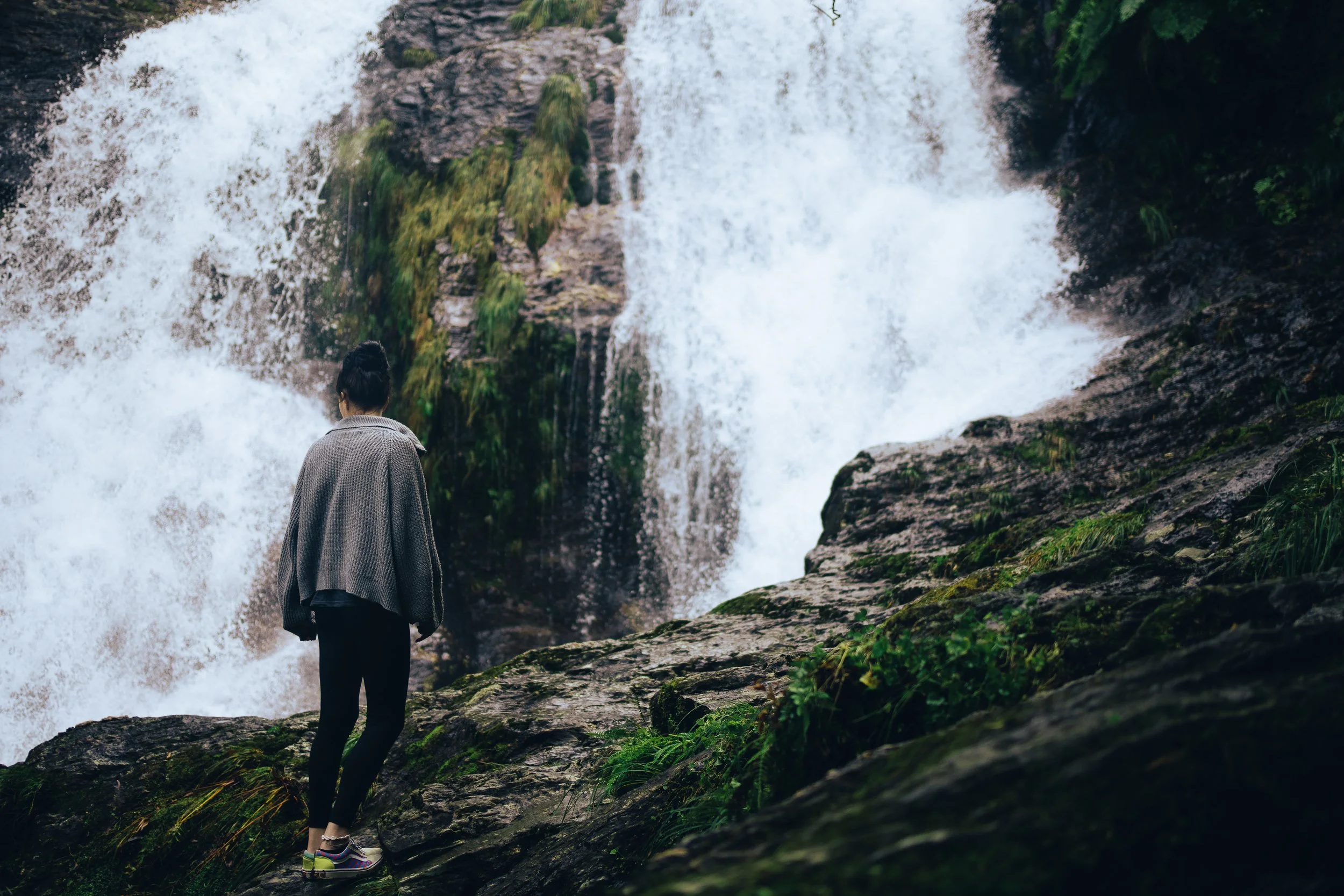 Eine Person steht auf einem Mossbewachsenen Felsen vor einem Wasserfall, trägt einen grauen Pullover und schwarze Hosen.