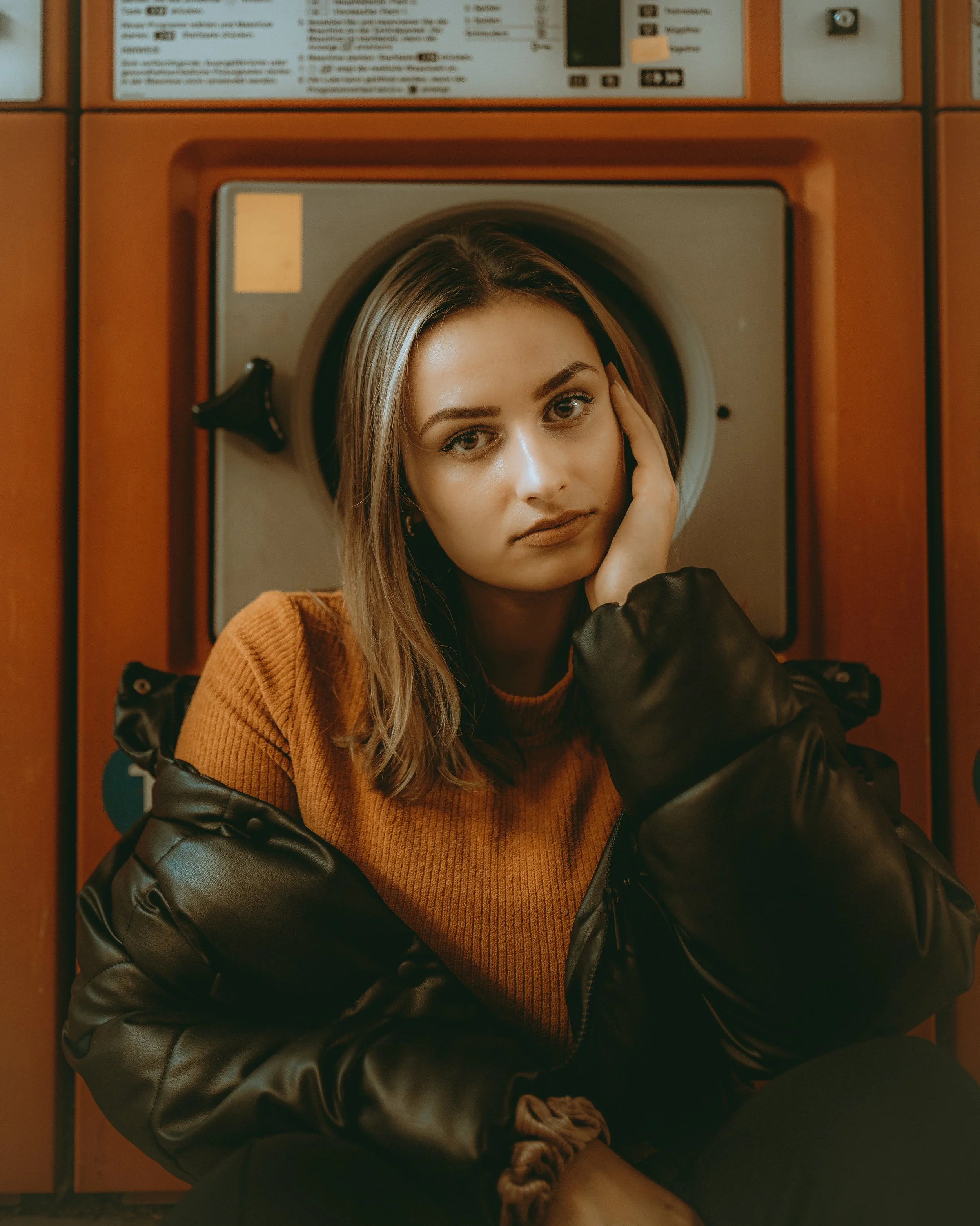 Junge Frau sitzt vor einer Waschmaschine, trägt einen braunen Pullover und einen schwarzen Pufferjacke, schaut nach vorne, lehnt ihren Kopf auf ihre Hand.