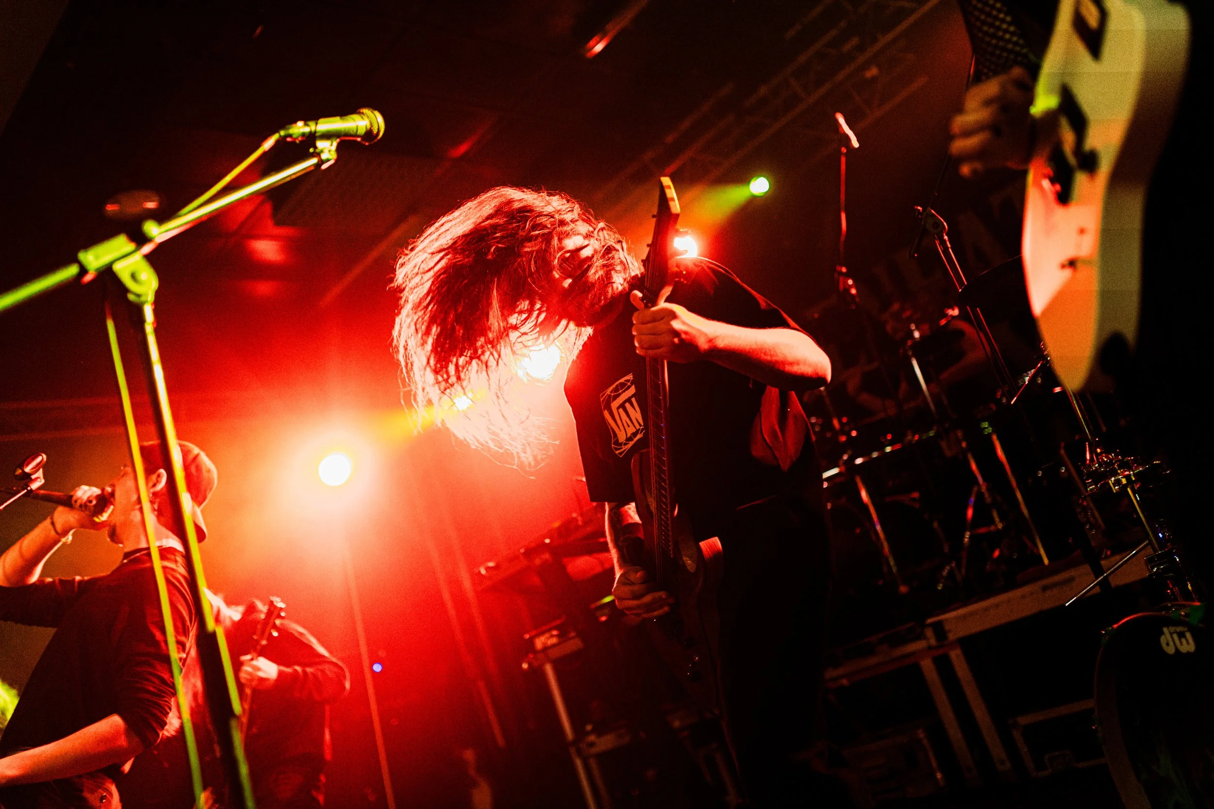 Zwei Musiker auf einer Bühne mit roter Beleuchtung, einer spielt Gitarre mit langem, lockigem Haar, die andere singt in ein Mikrofon, im Hintergrund Schlagzeug und weiteres Bühnen-Equipment