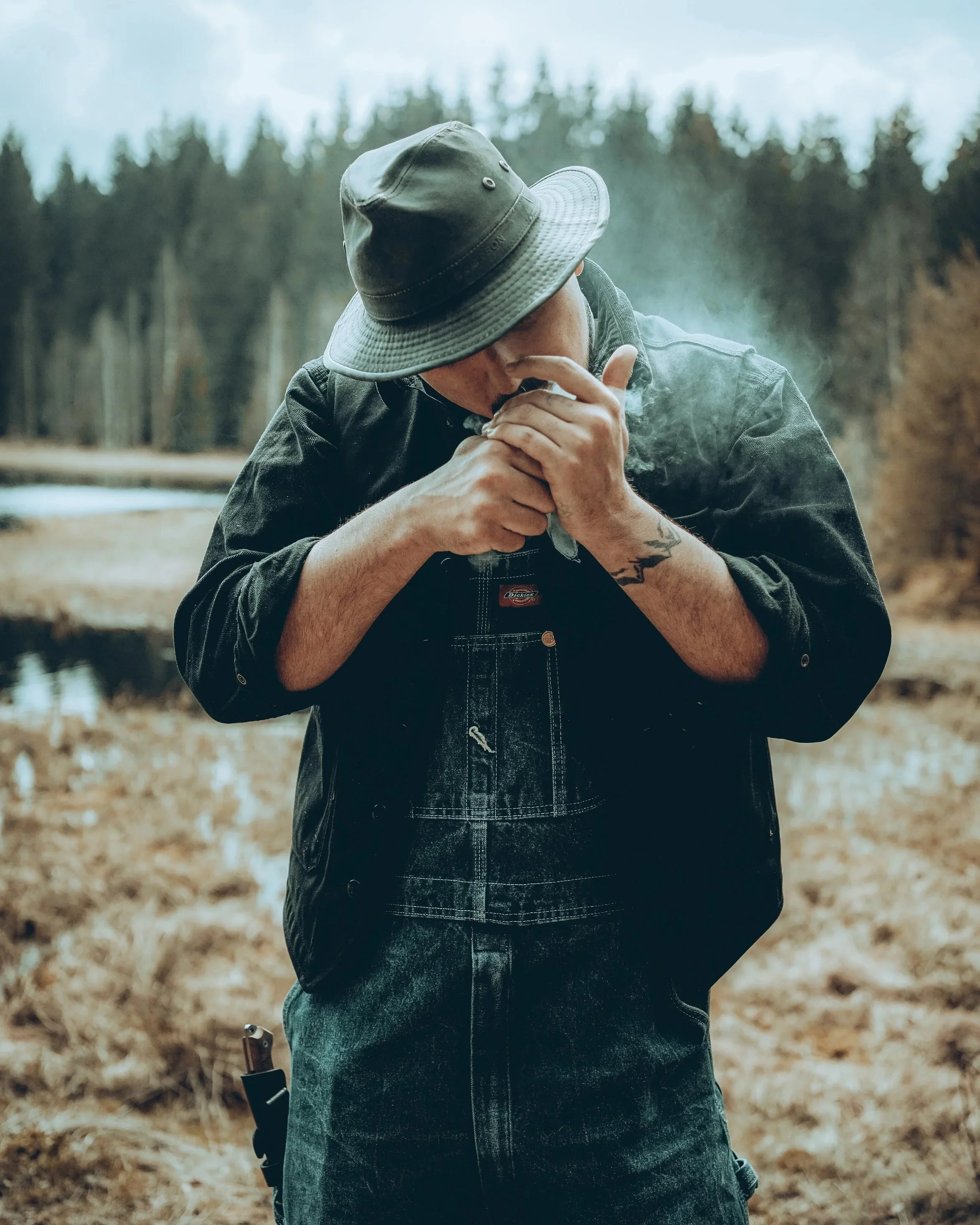 Ein Mann trägt einen Hut und eine dunkle Jacke, steht in einer natürlichen Umgebung und raucht.