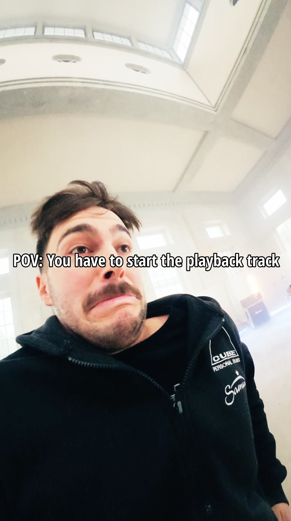 Ein junger Mann mit dunklem Haar und Bart steht in einem großen, hellen Raum mit hohen Fenstern. Er trägt eine schwarze Jacke mit einem Logo und Text. Der Text auf dem Bild lautet: "POV: You have to start the playback track".