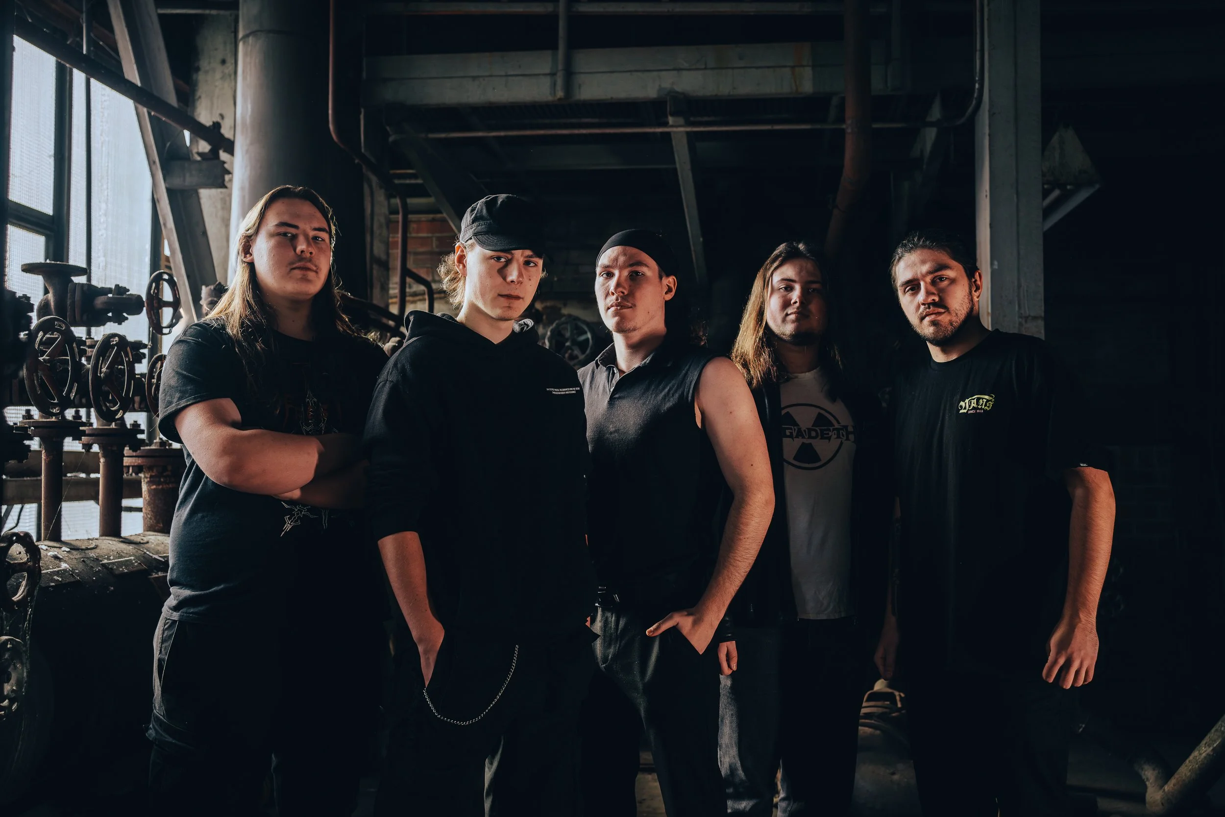 Fünf junge Männer posieren in einem dunklen, industriellen Raum mit Rohren und Ventilen im Hintergrund.